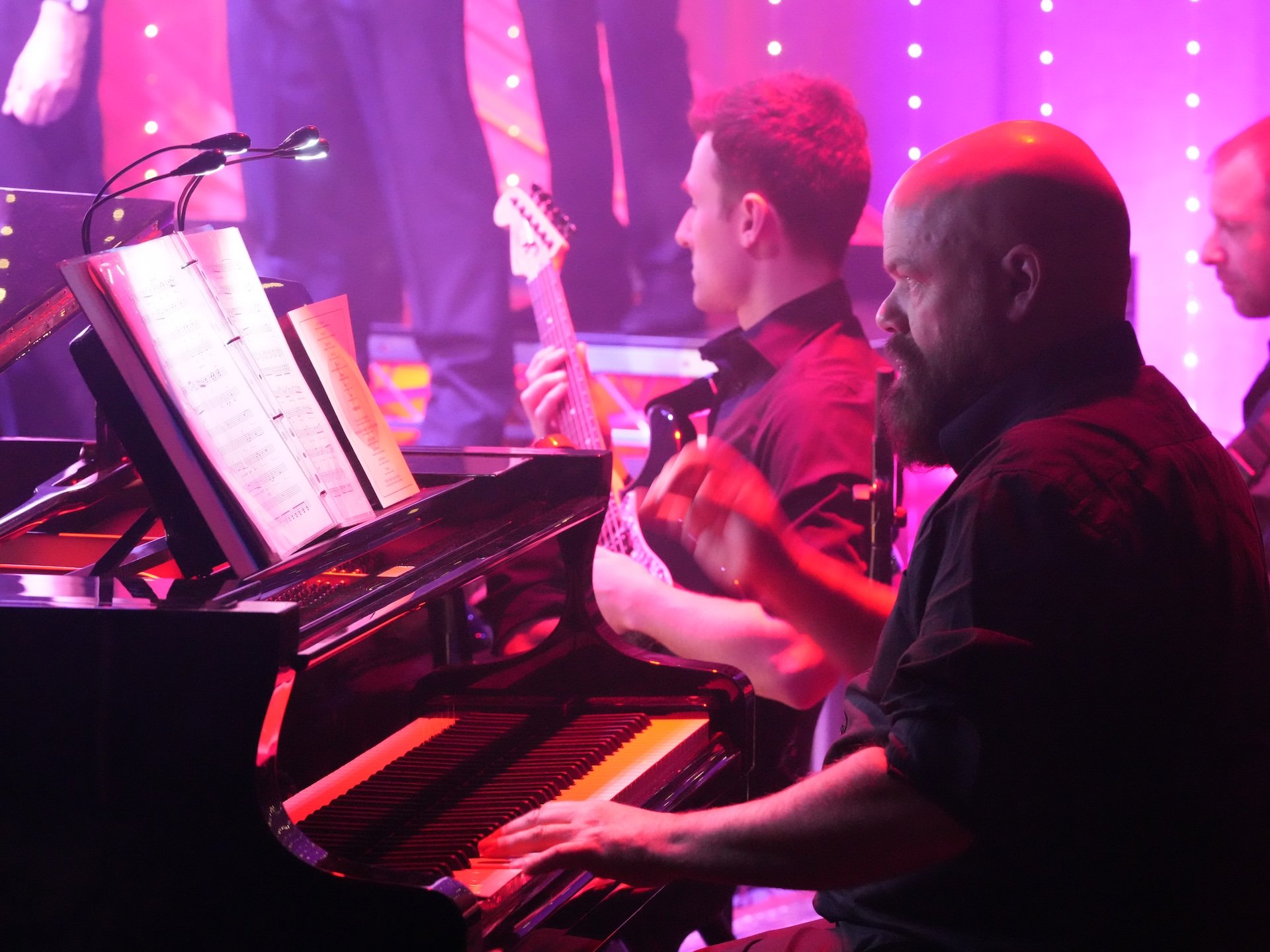 A group of musicians, including a pianist and guitar players, performing on stage under pink and purple lighting with sheet music on the piano.