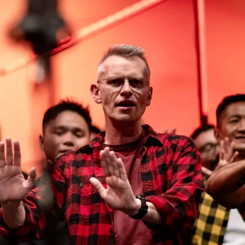 A man with glasses and short hair wearing a red and black plaid shirt at a gathering, with several other people in the background, some with hands raised.