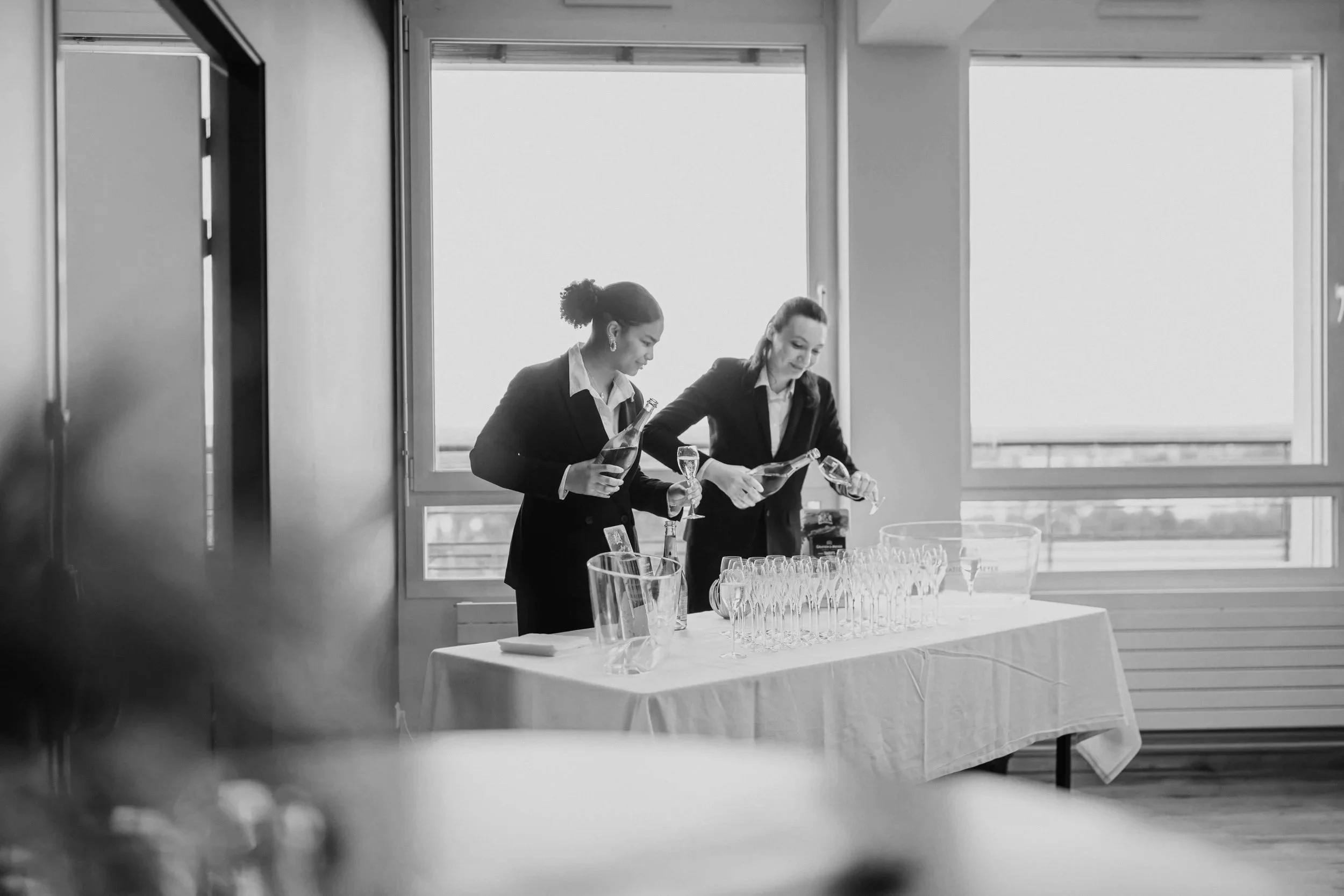 Two women in professional attire preparing drinks at a table with glasses and bottles, near large windows in a bright room.