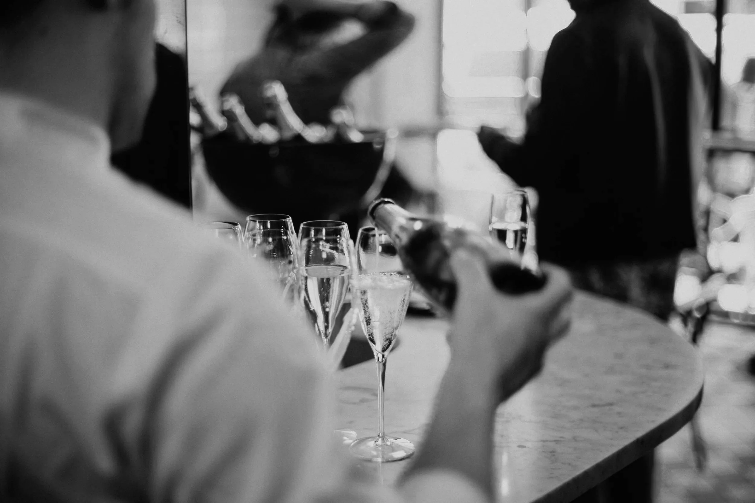 A person pouring champagne into a flute at a celebration, with several filled champagne glasses on the table, in a black and white photo.