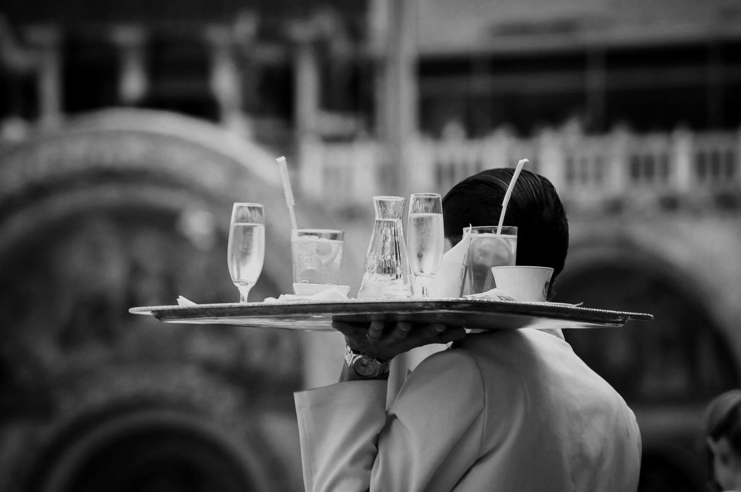 A waiter carrying a tray with drinks on his shoulder; the drinks include a glass of water, a flute of champagne, and a glass with a straw, in black and white.