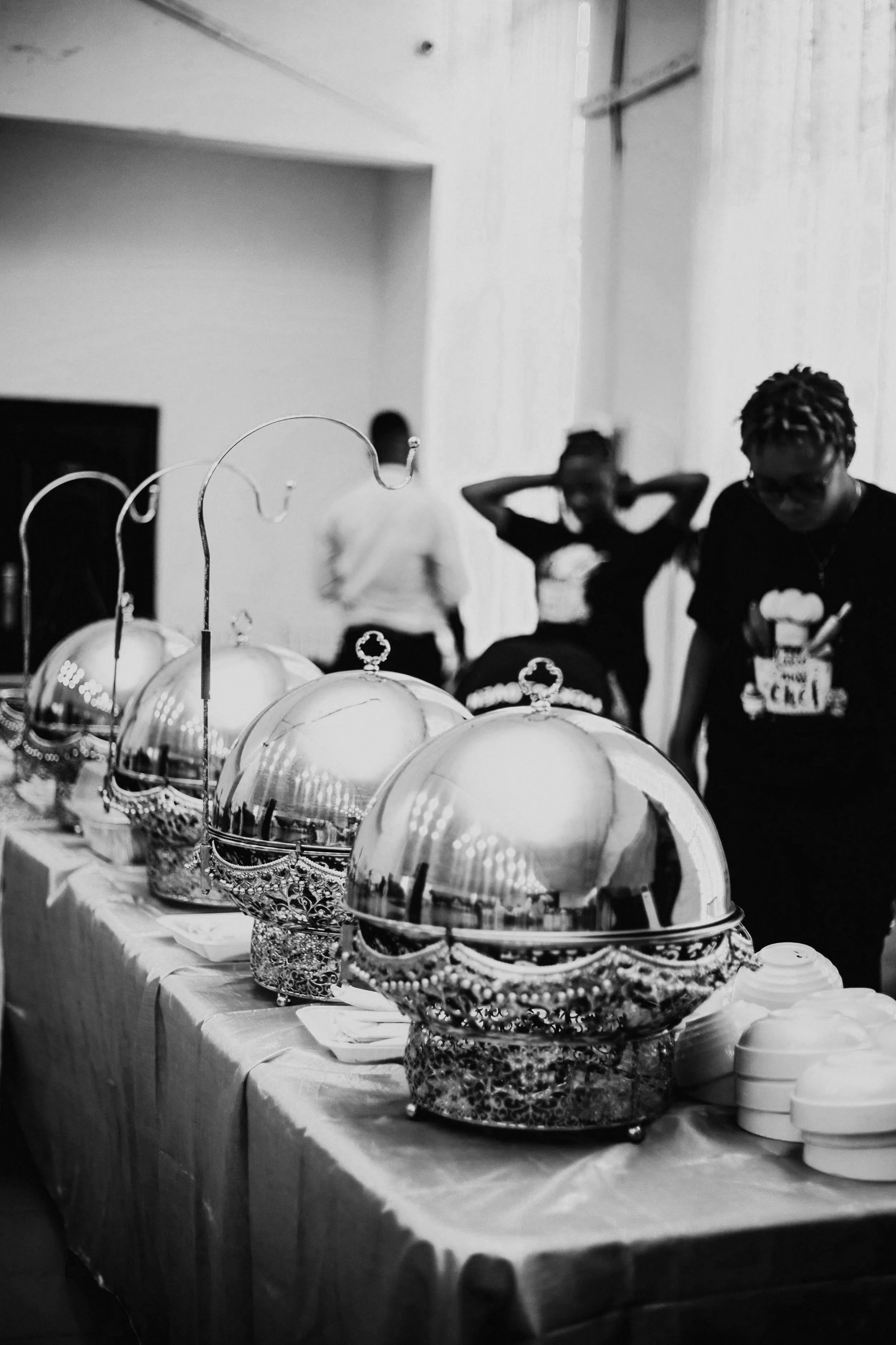 A row of ornate, silver chafing dishes with domed lids on a table, with people in the background, one with hands behind head and another serving food.