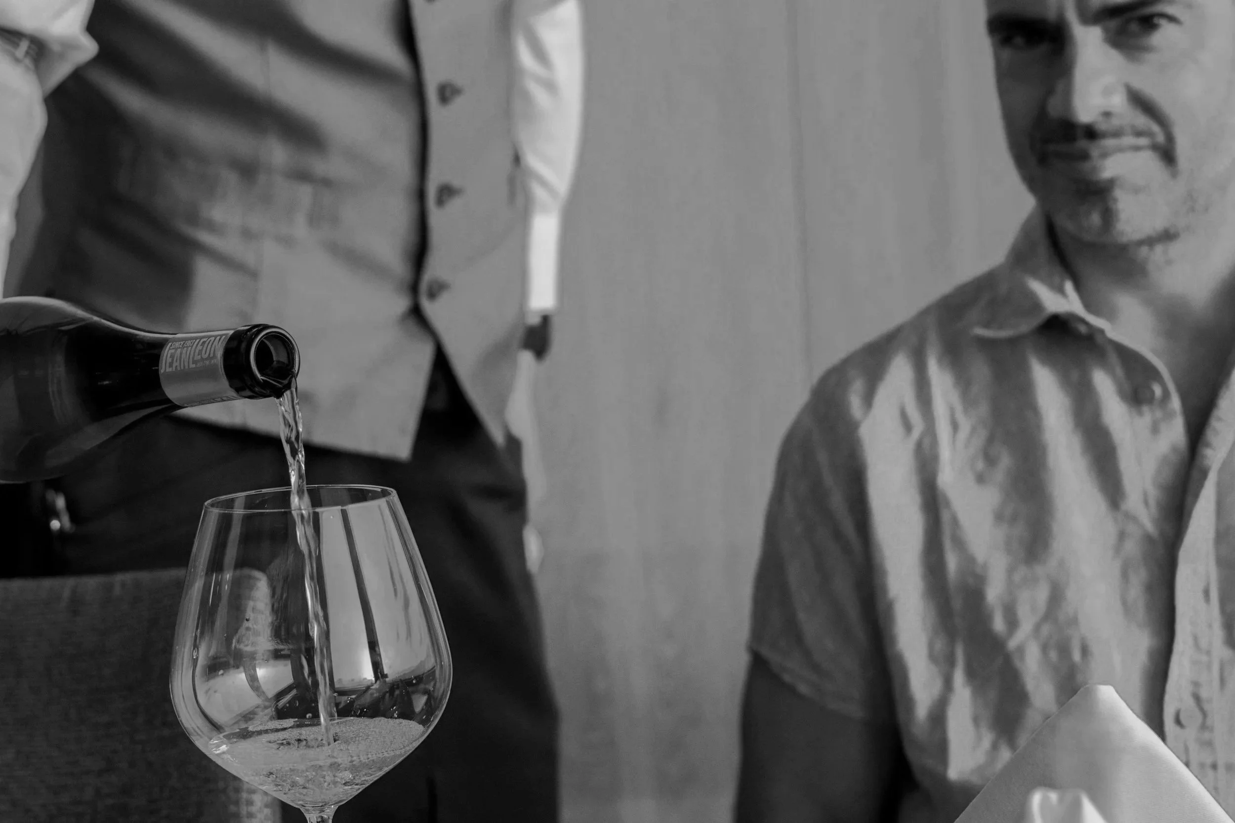 A man in a light-colored shirt sitting at a table with a white napkin, a bottle pouring wine into a glass, while another person in a vest stands nearby. The scene is in black and white.