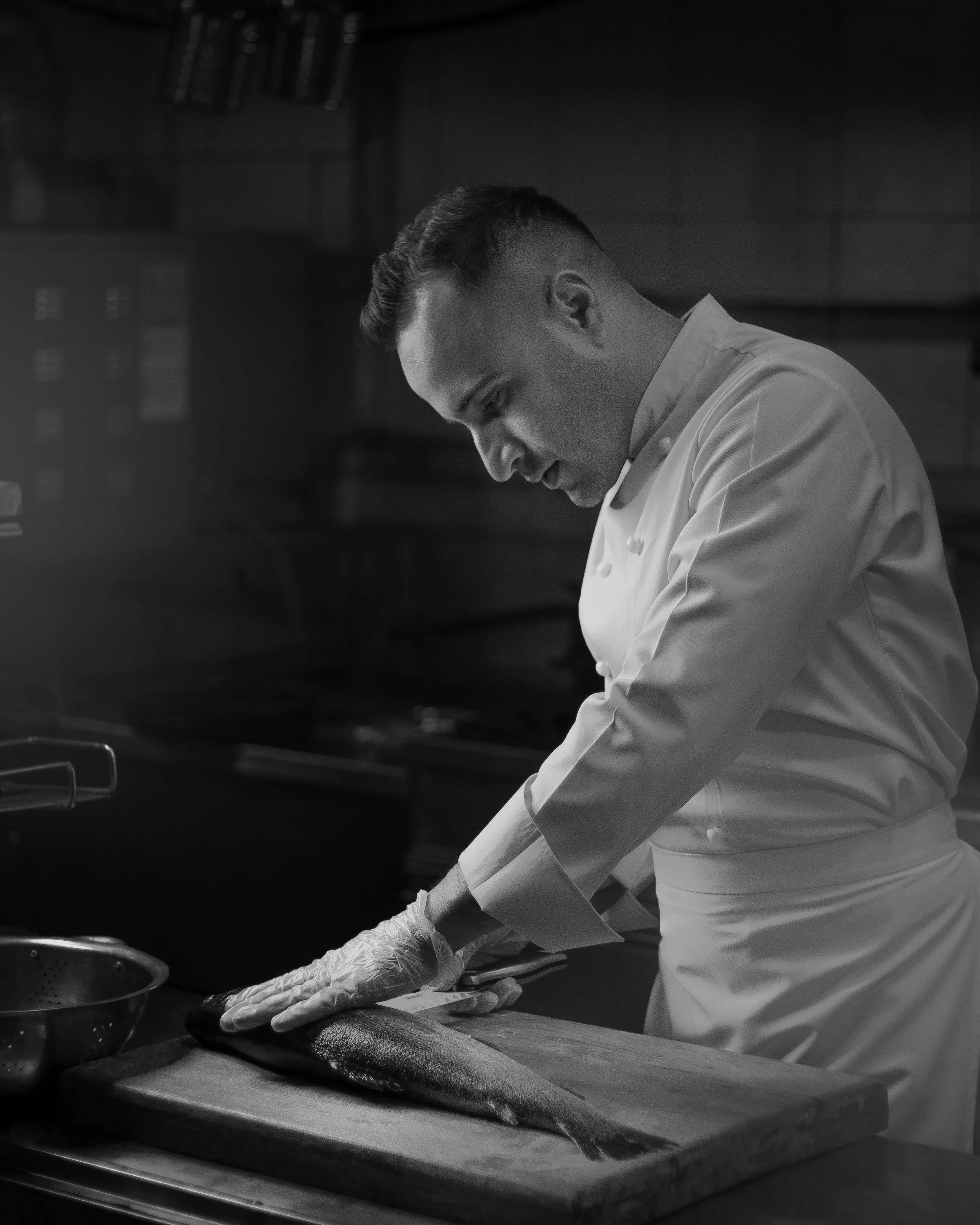 Chef in a white uniform and gloves preparing a fish on a wooden cutting board in a kitchen.