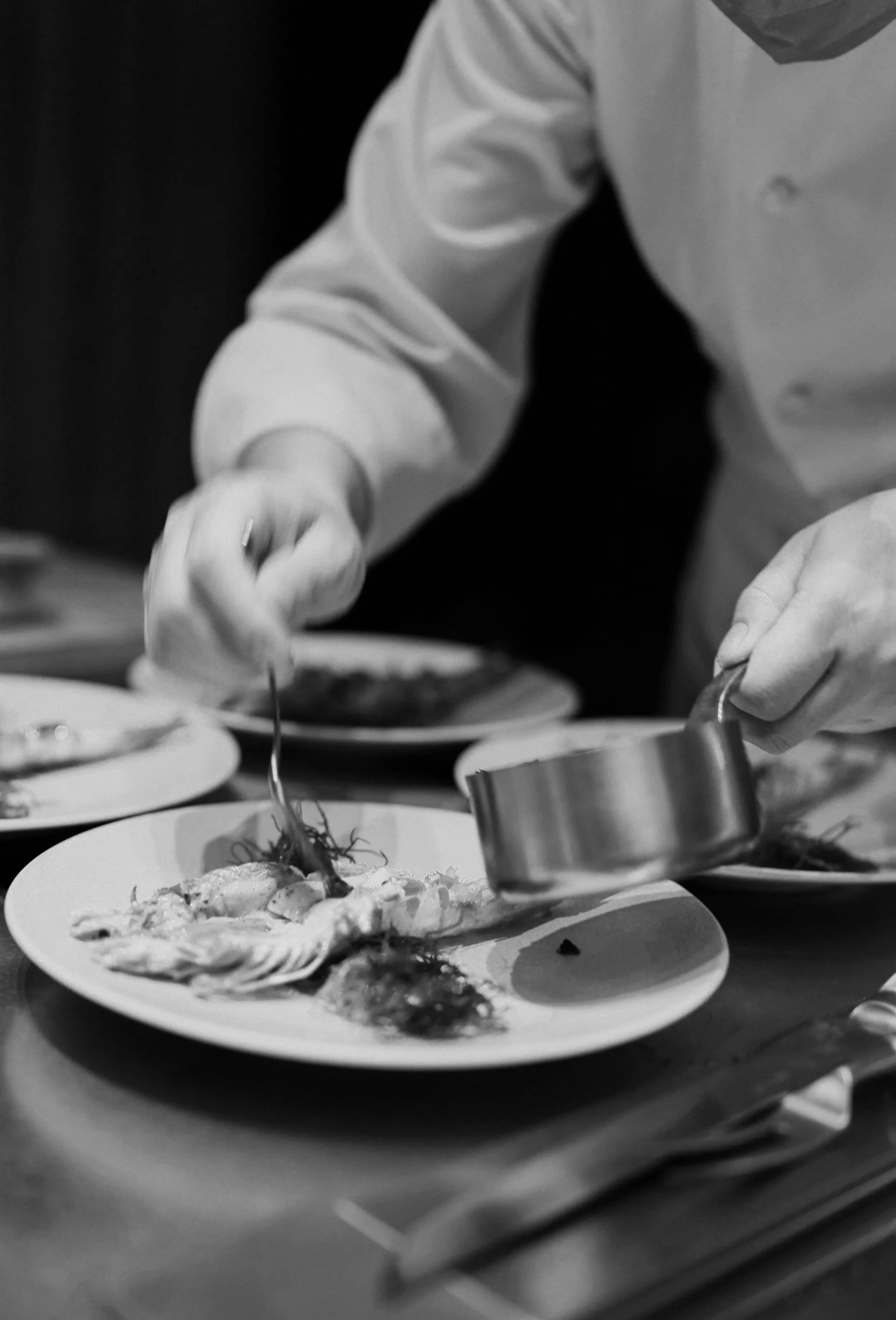 Chef in a white coat elegantly plating a fish dish garnished with herbs and sauce in a professional kitchen.