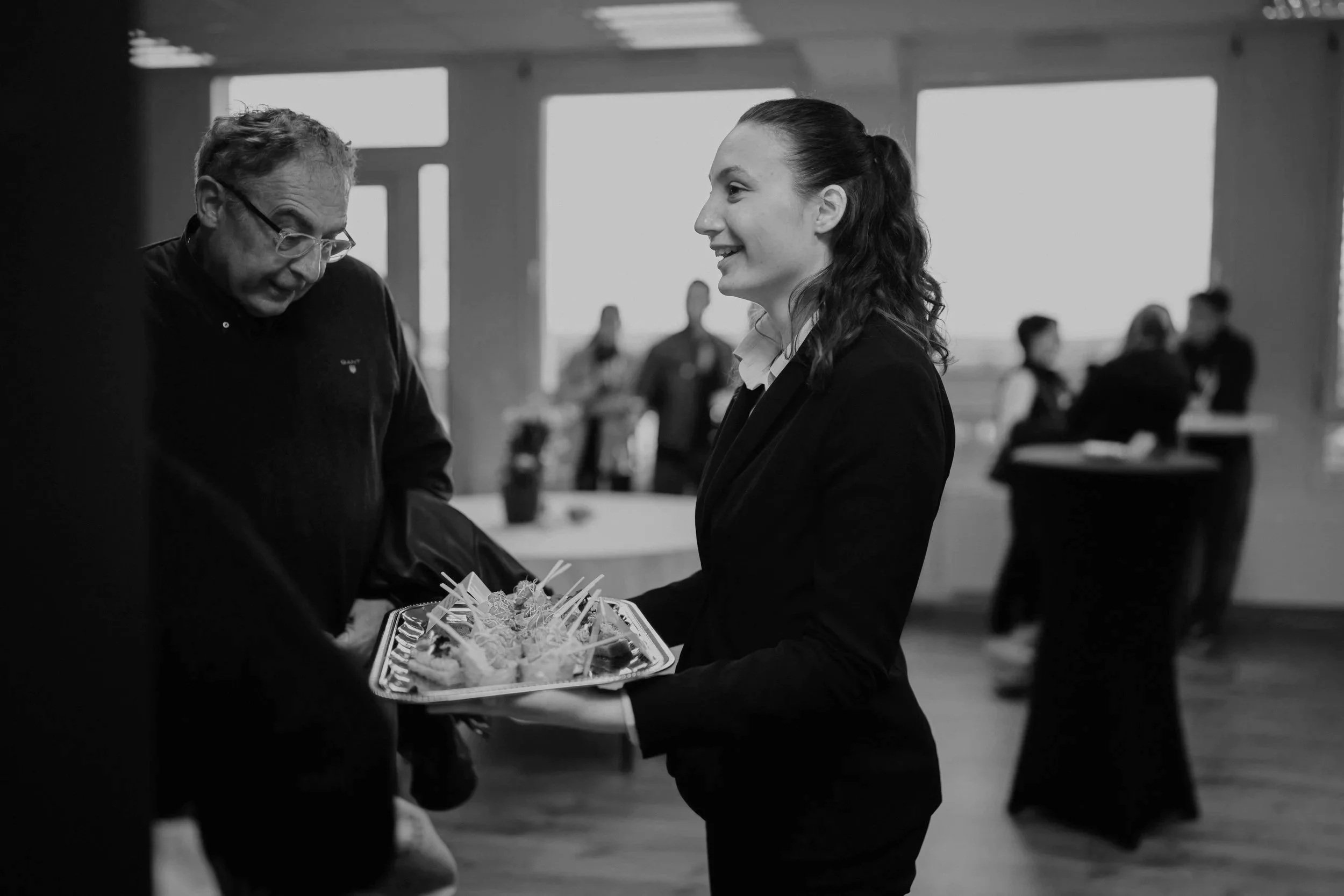 A woman presenting a platter of appetizers or snacks to an older man at an indoor social event.