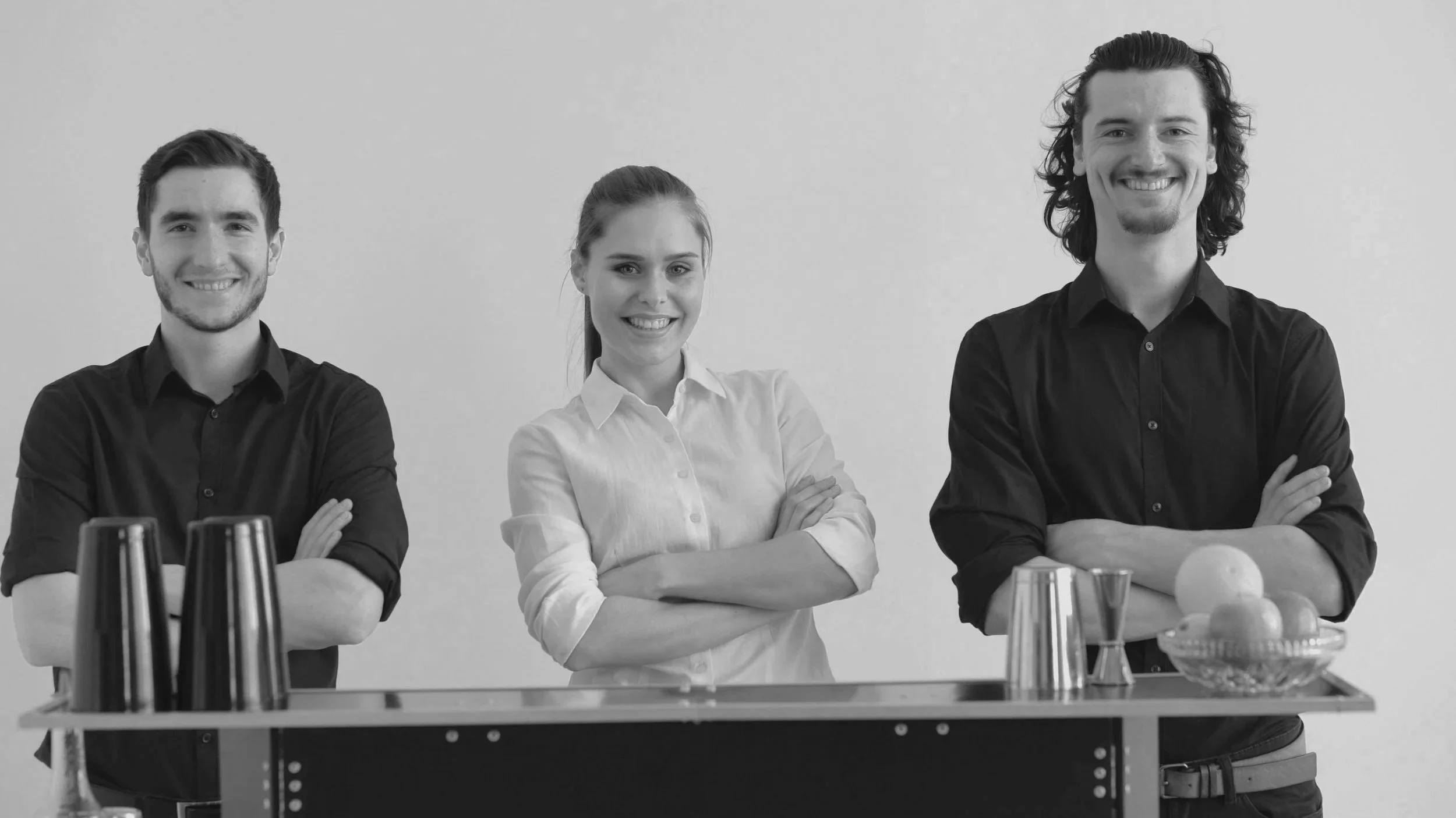 Three people standing behind a bar with cups, a bowl of fruit, and drinks, smiling with arms crossed.