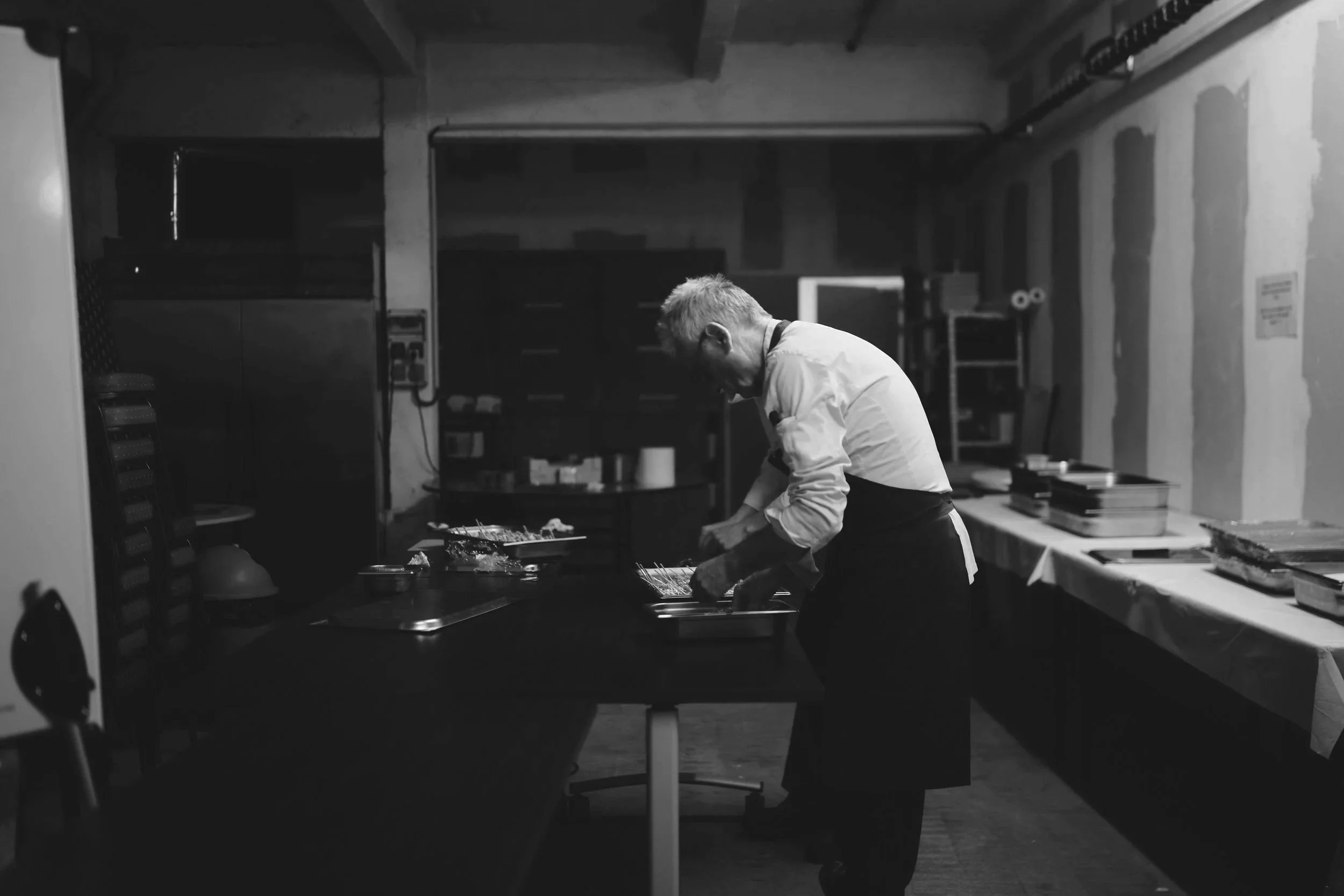 A chef preparing food in a kitchen, wearing a white chef coat and black apron, with a workspace cluttered with trays and kitchen supplies.