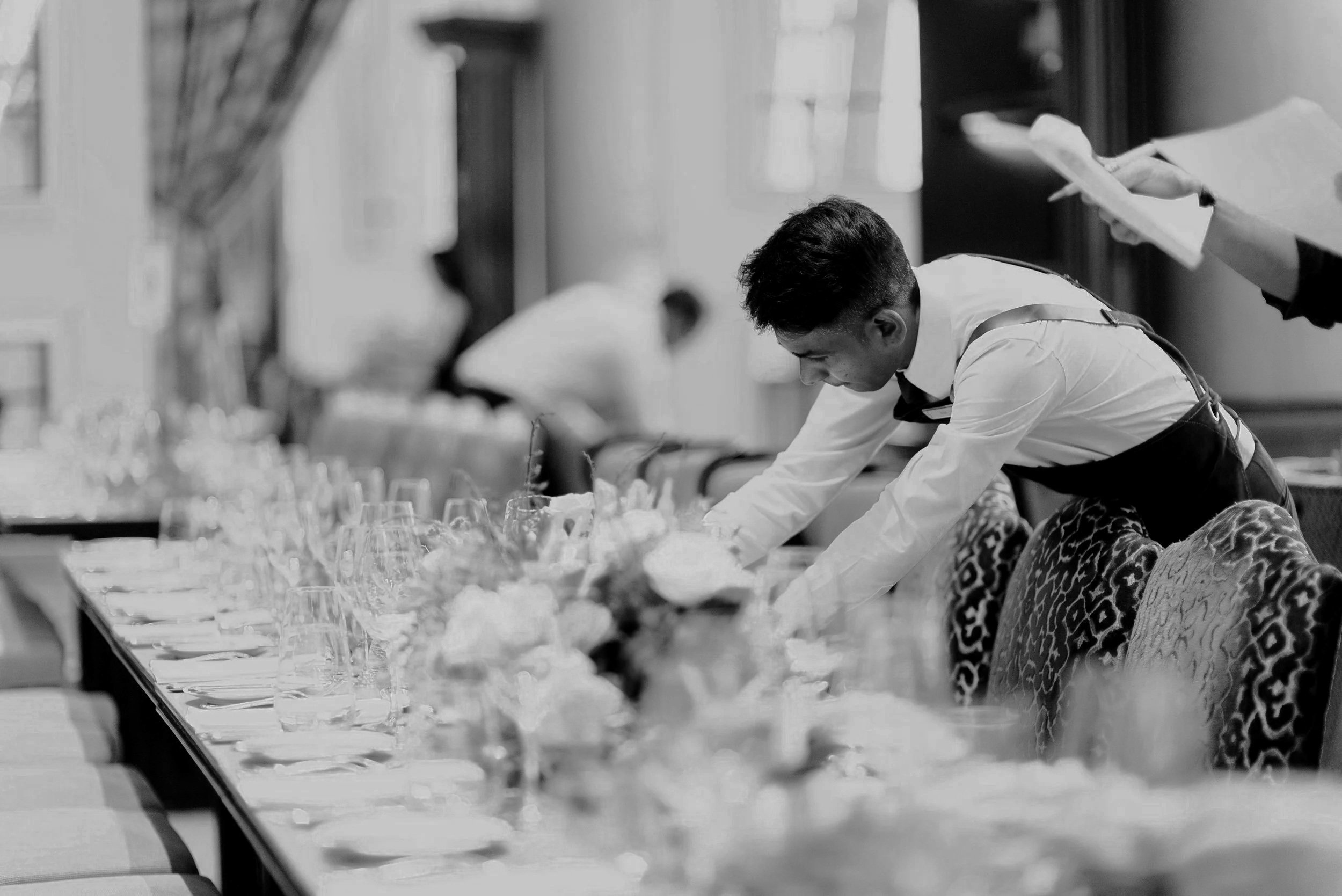 A young man in formal attire setting a banquet table with glasses and floral centerpieces in a decorated dining room.
