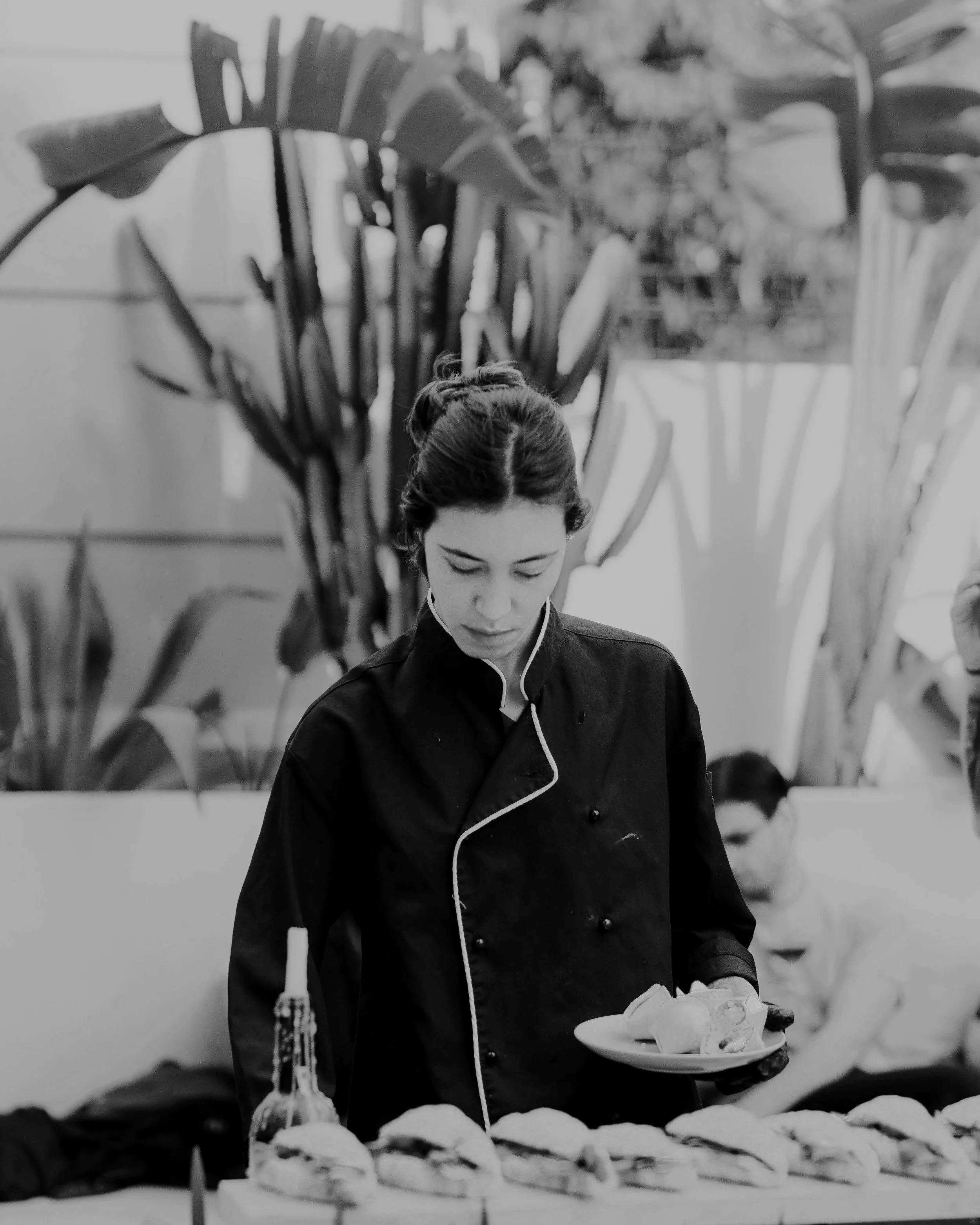 A female chef in a black uniform serving food at a buffet, with large leaf plants in the background.