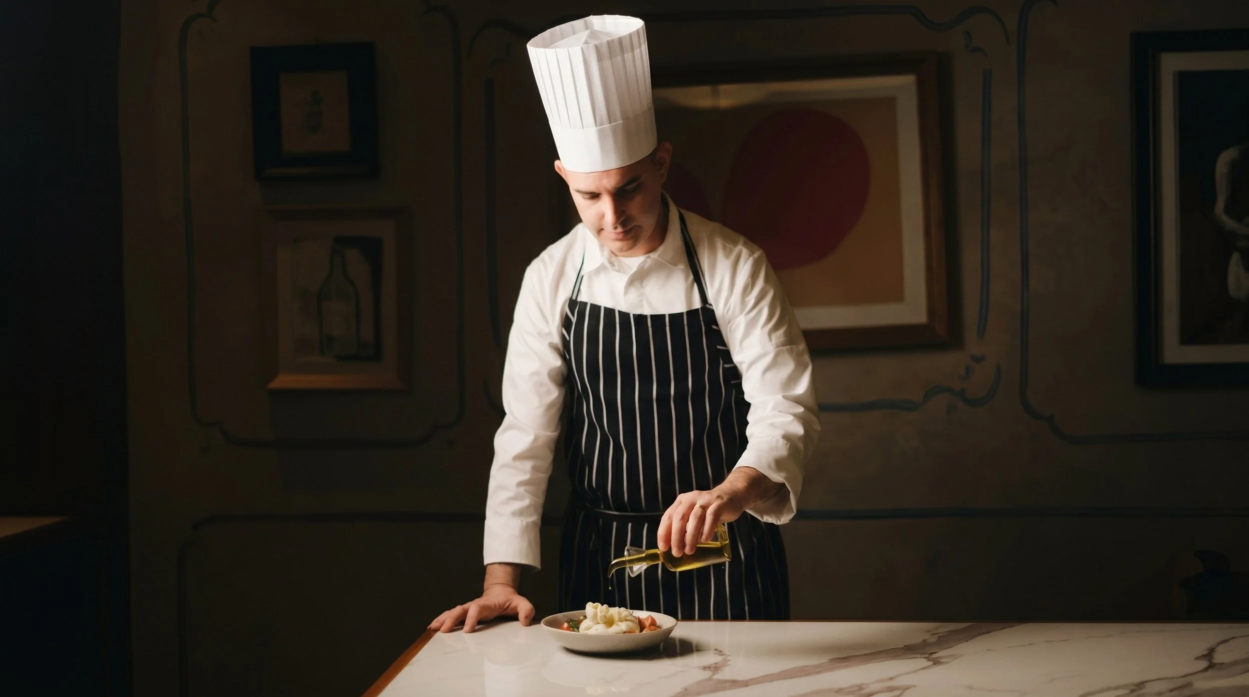 A chef in a white uniform and black-striped apron, wearing a tall white chef's hat, is pouring olive oil onto a dish on a white marble table.