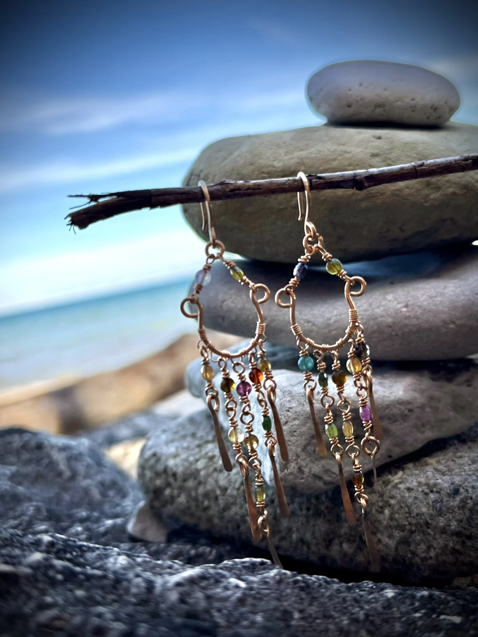 Colorful beaded earrings hanging from a wooden stick, with layered stones and a beach in the background.