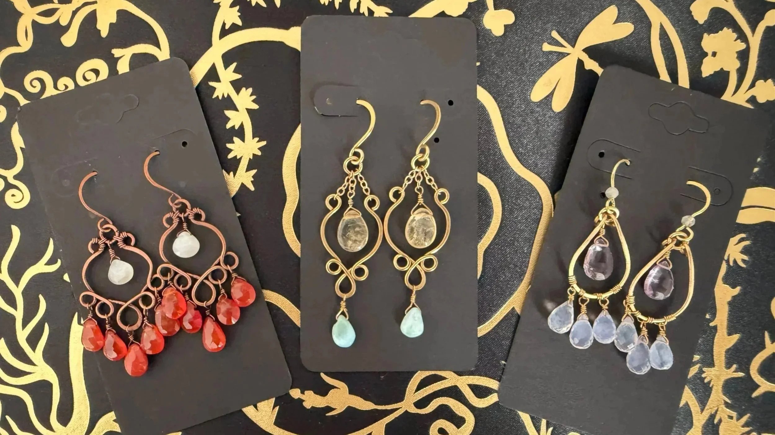 Three pairs of dangle earrings with gemstone beads on black display cards, set against a black and gold decorative background.