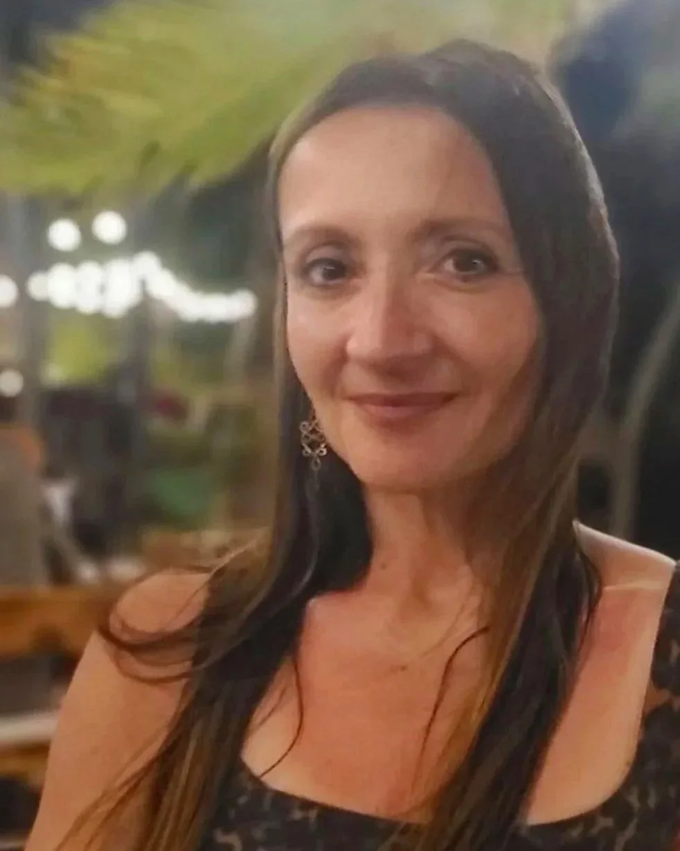 A woman with long dark hair and earrings smiling in a dimly lit indoor setting.