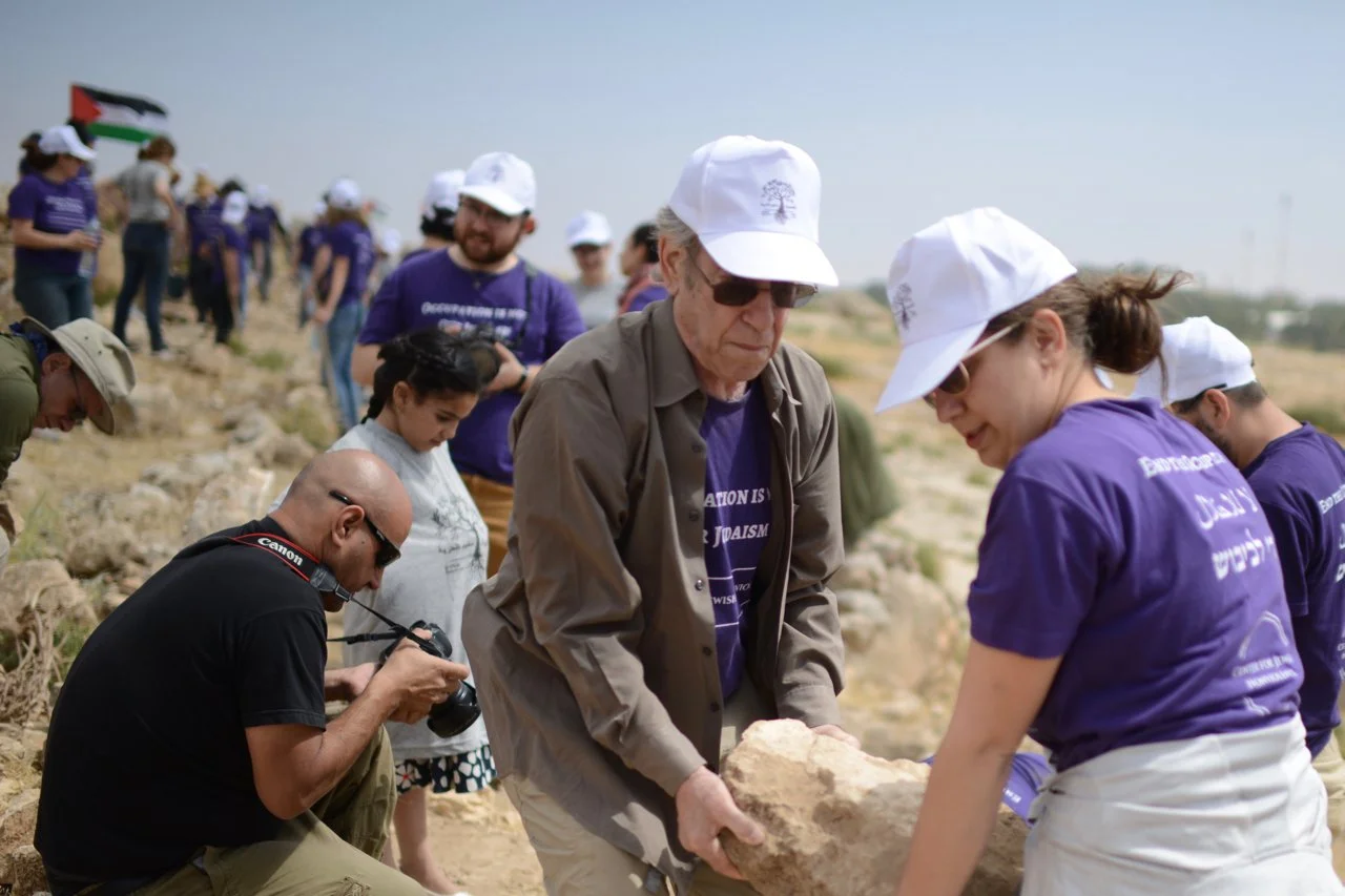 A group of people on a rocky outdoor terrain, some wearing purple T-shirts and white hats, participating in a collective activity involving a large rock. One person is taking photos, others are examining or moving the rock. An Egyptian flag is visibl