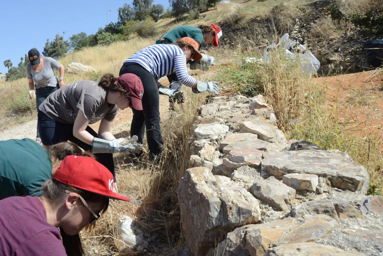 Group of people working outdoors to preserve or restore a stone wall on a hillside.