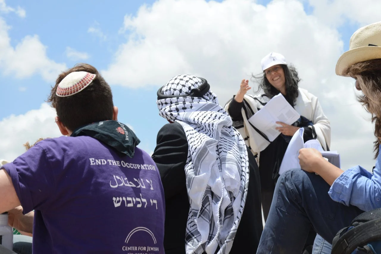 A group of people gathered outdoors, some wearing traditional Middle Eastern attire and head coverings, listening to a woman speaking while holding papers, with a blue sky and clouds in the background.
