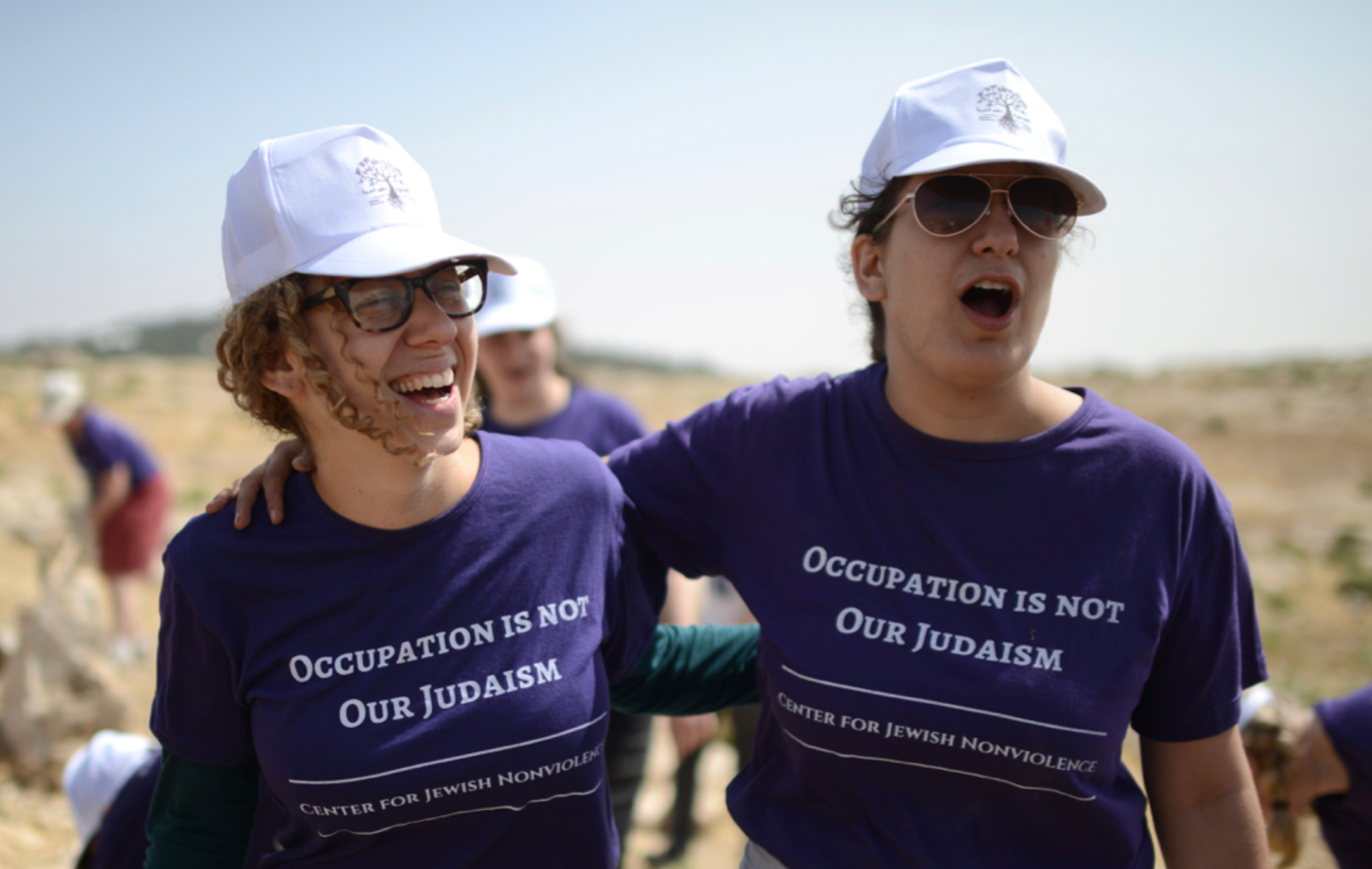 Two women wearing white caps and matching purple T-shirts participating in an outdoor event. They are smiling and appear to be happy, with one woman having her arm around the other.