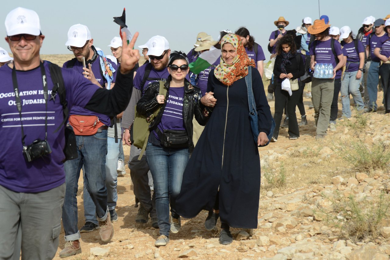 A group of tourists walking outdoors in a desert-like area, many wearing purple shirts that read 'Occupation is not Judaism' and holding backpacks. A woman in black attire and headscarf is holding hands with another woman in sunglasses and a black ja