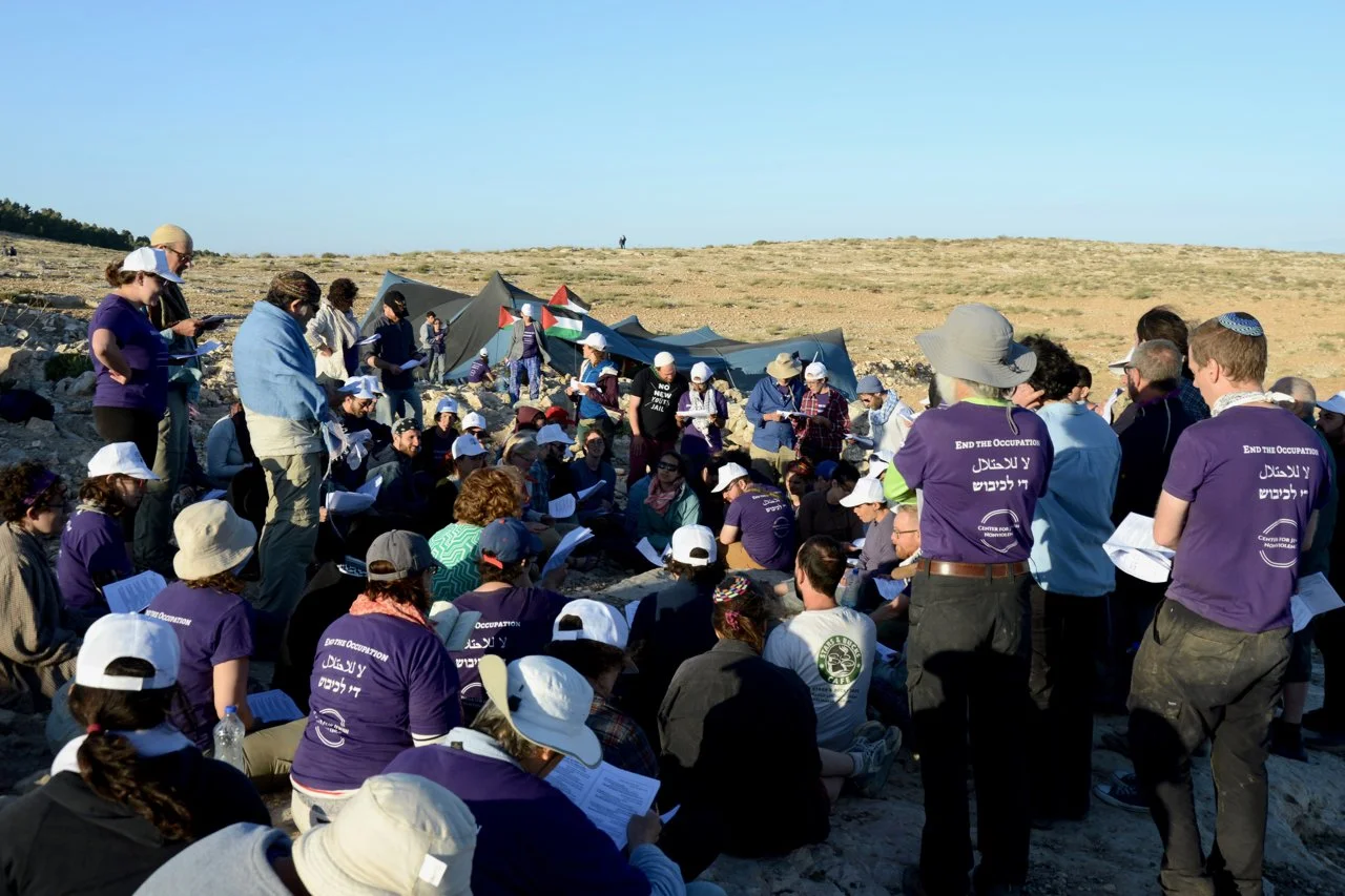 A large group of people gathered outdoors in a desert landscape, participating in a protest or demonstration, with some holding papers and wearing purple shirts with slogans.