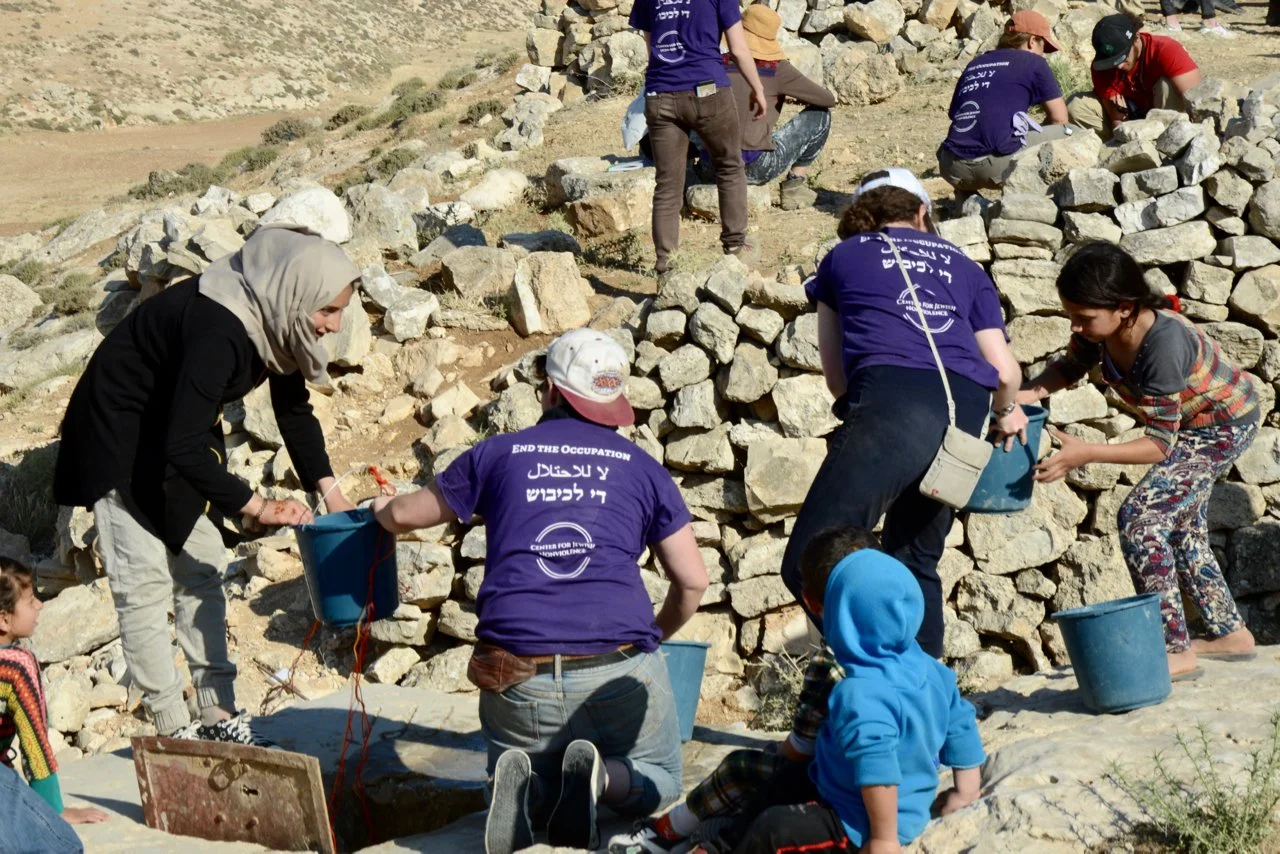 People working on an archaeological site with stone ruins in a desert landscape, some wearing purple shirts with writing and others assisting each other.