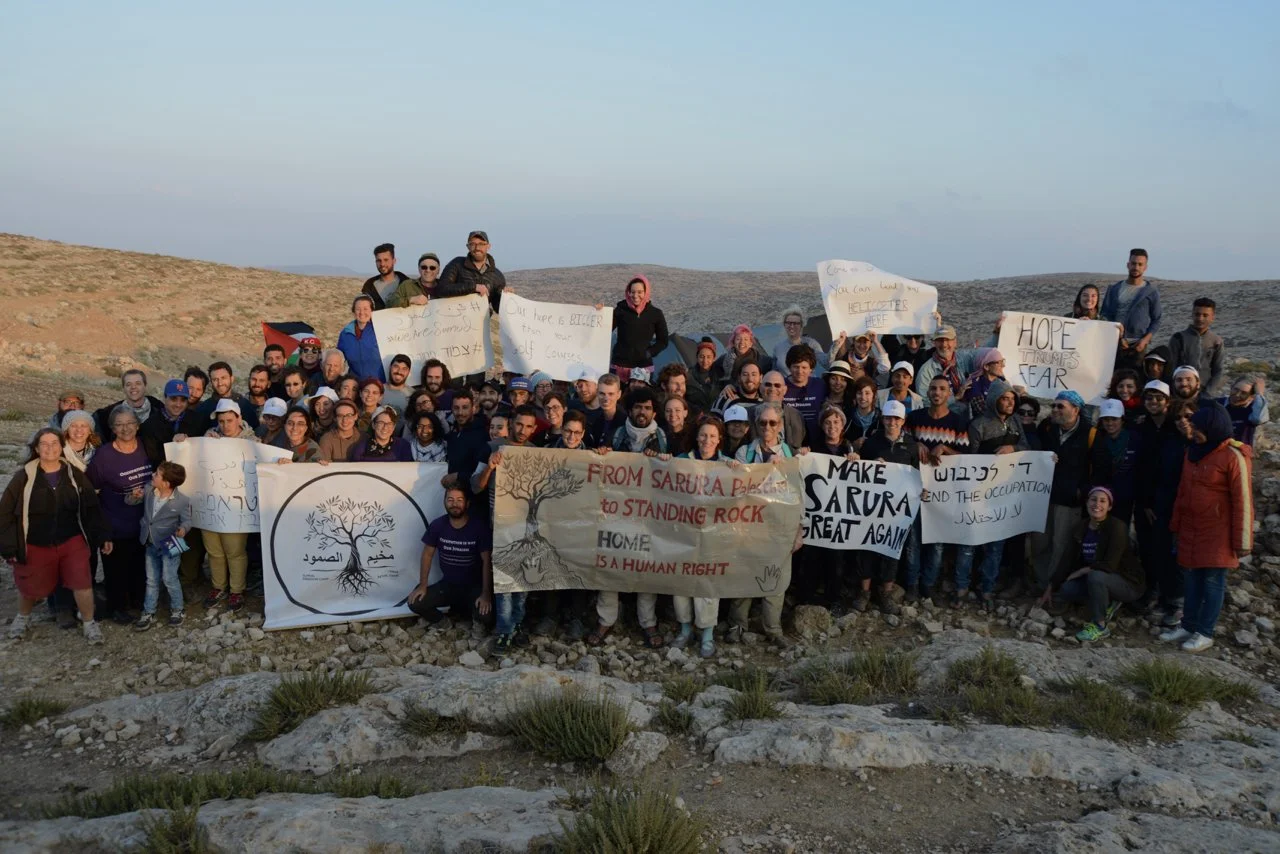 Group of people holding protest signs and banners in a rocky outdoor area, with hills in the background.