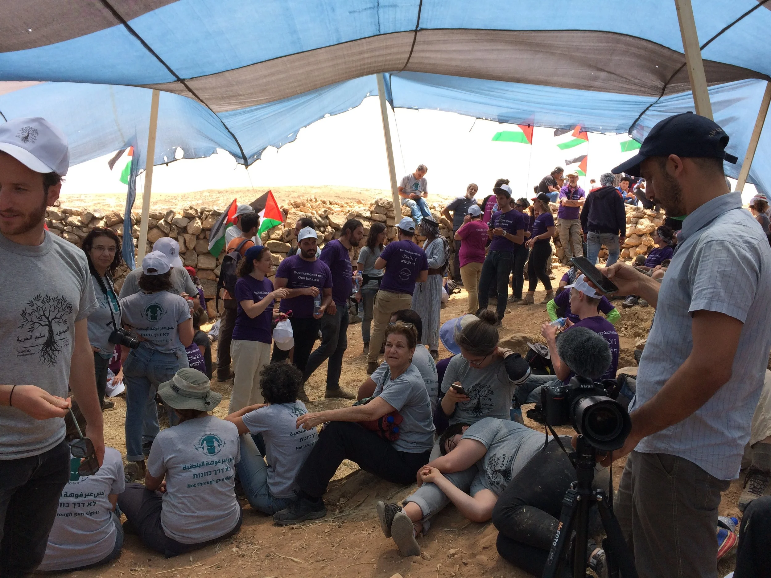 A large group of people gathered outdoors under a makeshift canopy made of fabric and wooden poles. Some are standing, some are sitting or crouching on the ground, and several are holding flags or looking at their phones. The crowd appears to include
