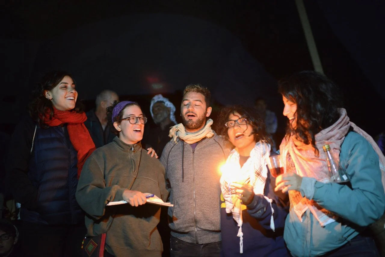 Group of five people singing and smiling, one holding a candle, in a dark setting.