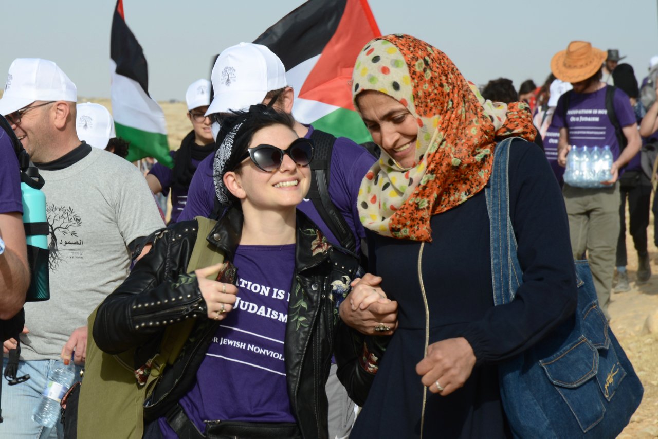 Two women smiling and holding hands at a rally or protest, with other people in the background holding flags and water bottles, in a desert area.