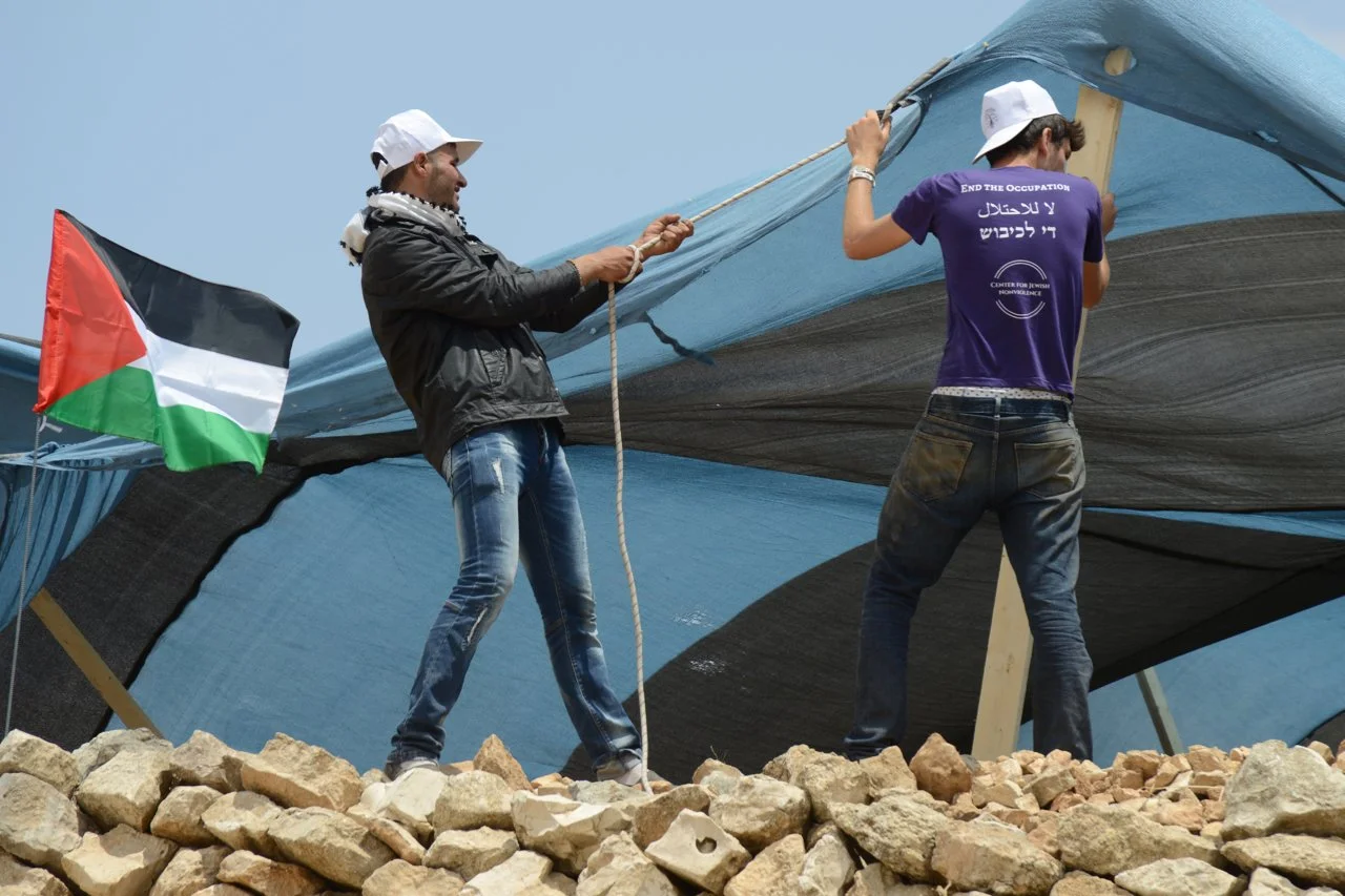 Two people setting up a large black and blue tent outdoors on rocky ground. One person is holding a rope, and the other is securing the tent. An Palestinian flag is visible on the left side of the image.
