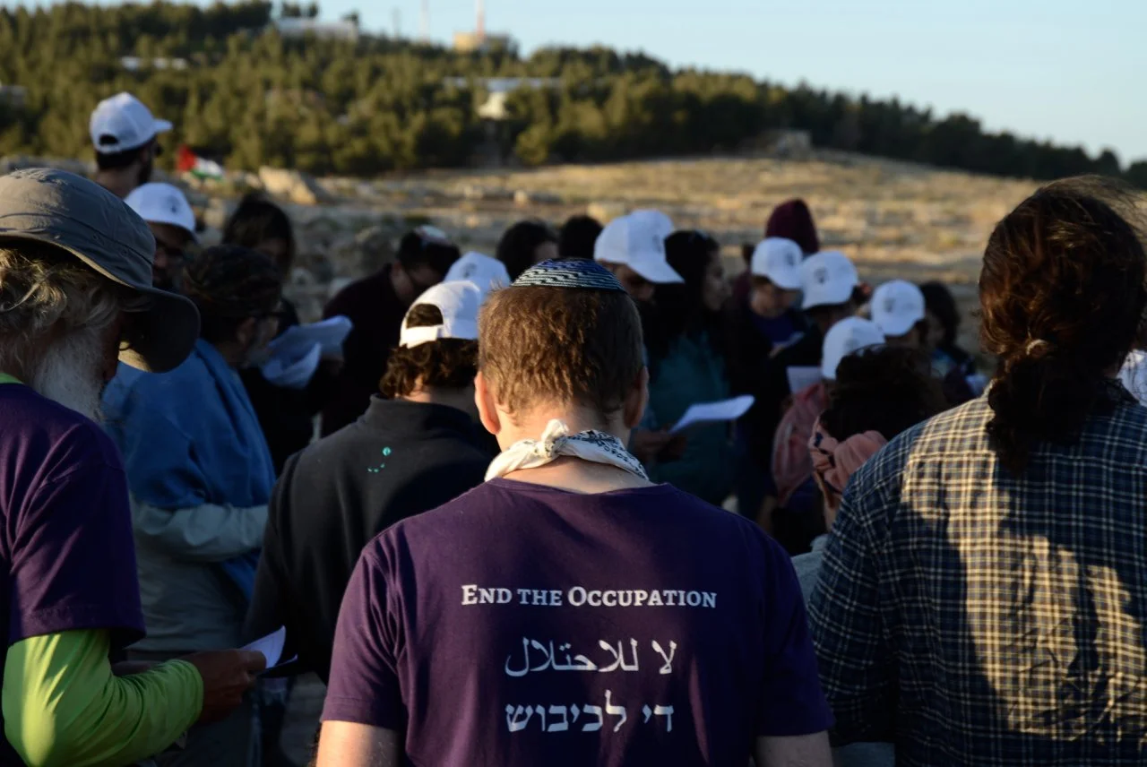 A group of people gathered outdoors during daytime, some holding papers, with many wearing white caps. One person in the foreground is wearing a purple shirt with the message 'End the Occupation' in English and Arabic. The background features trees a