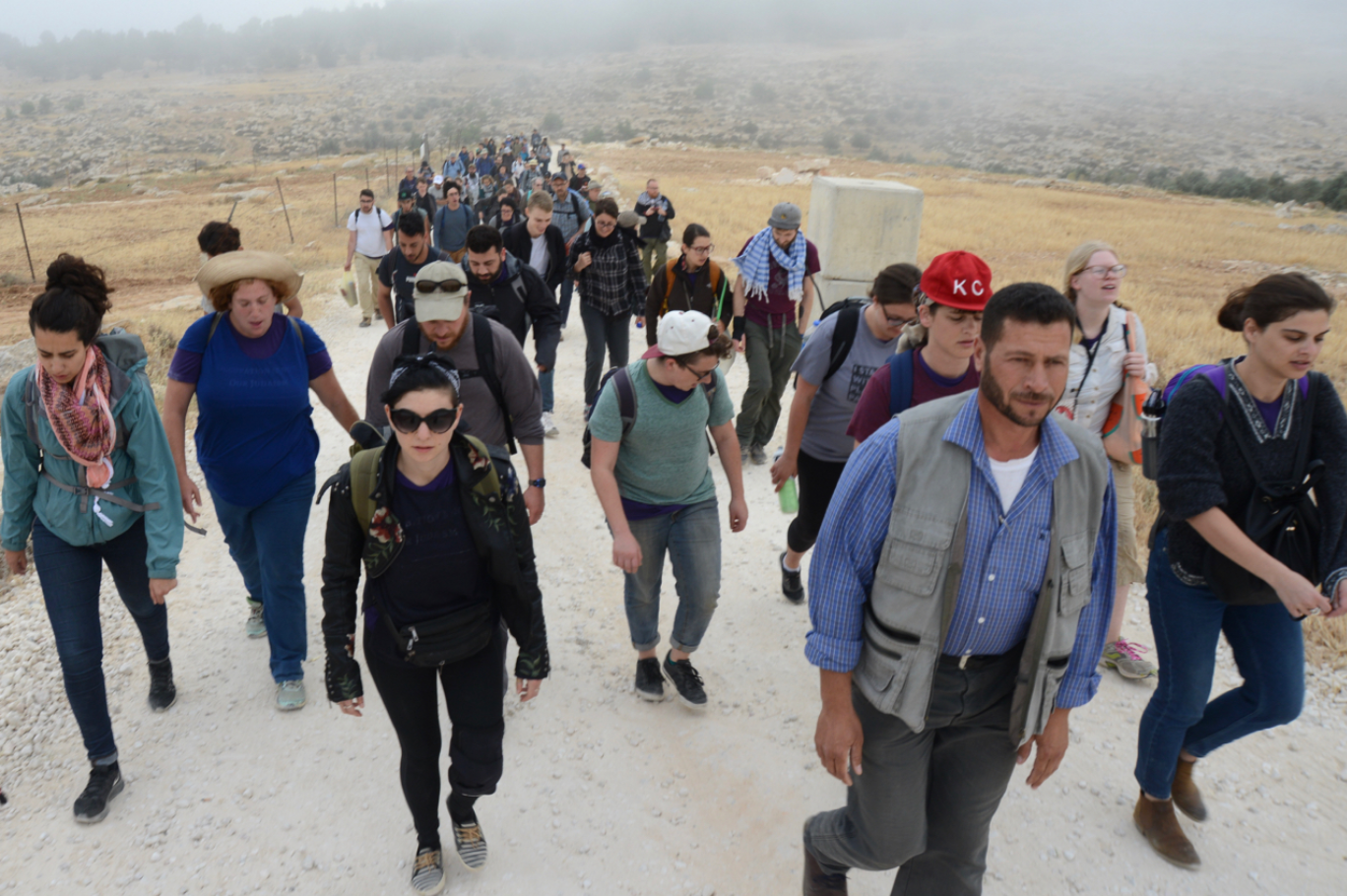Group of people hiking on a dirt trail in a dry, grassy landscape with hills in the background.