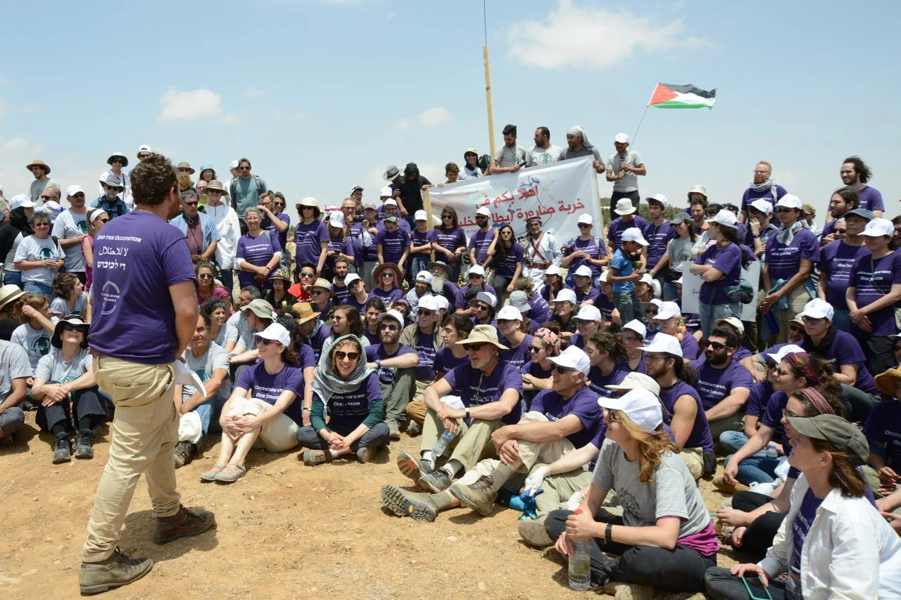 A large group of people gathered outdoors on a sunny day, many wearing purple shirts and hats, some holding a Palestinian flag, participating in a protest or rally with a speaker addressing the crowd.