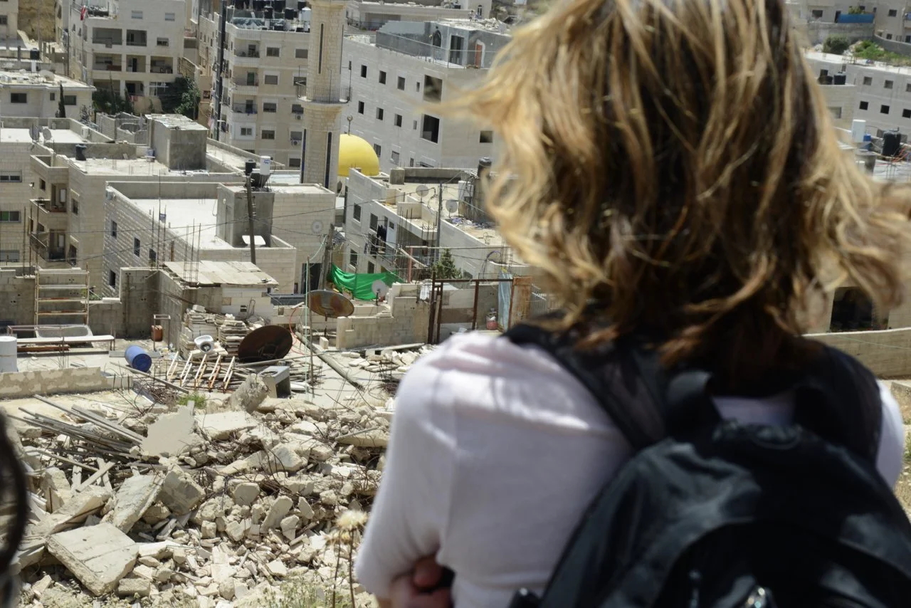 Person with curly blonde hair wearing a white shirt and black backpack looking over a damaged building in an urban area with debris and rubble.