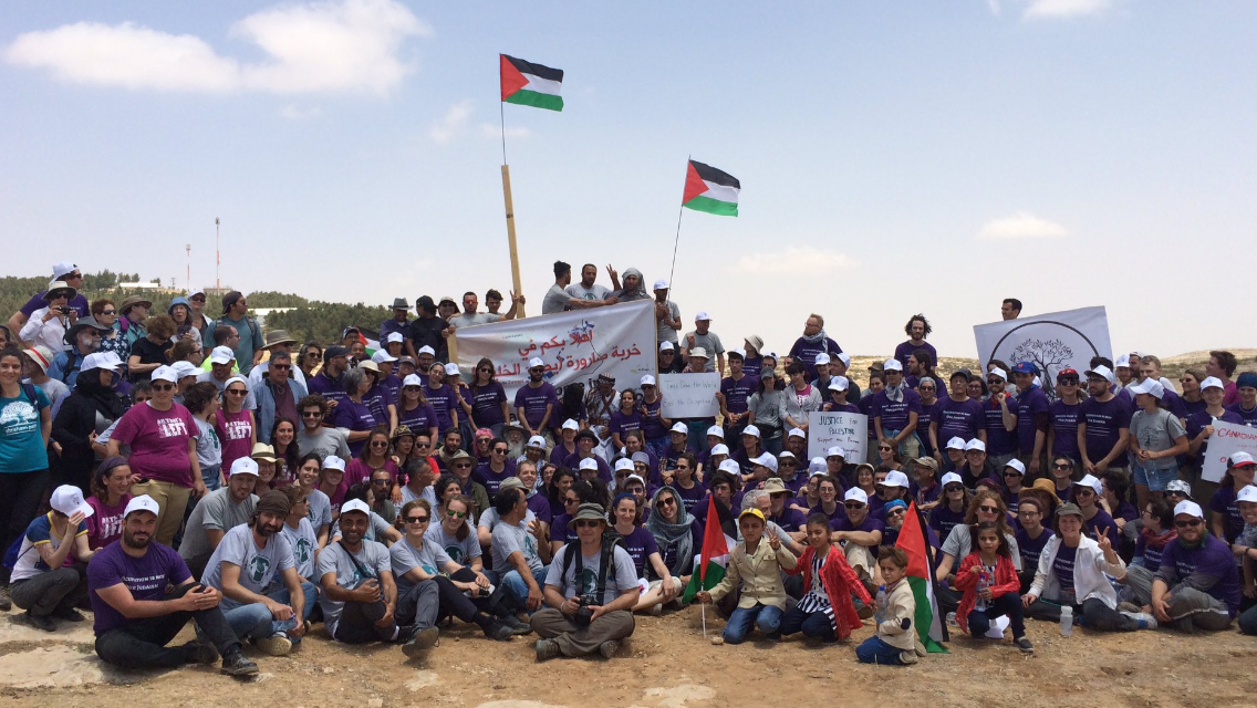 Large group of people gathered outdoors, holding Palestinian flags and signs, participating in a demonstration or protest.