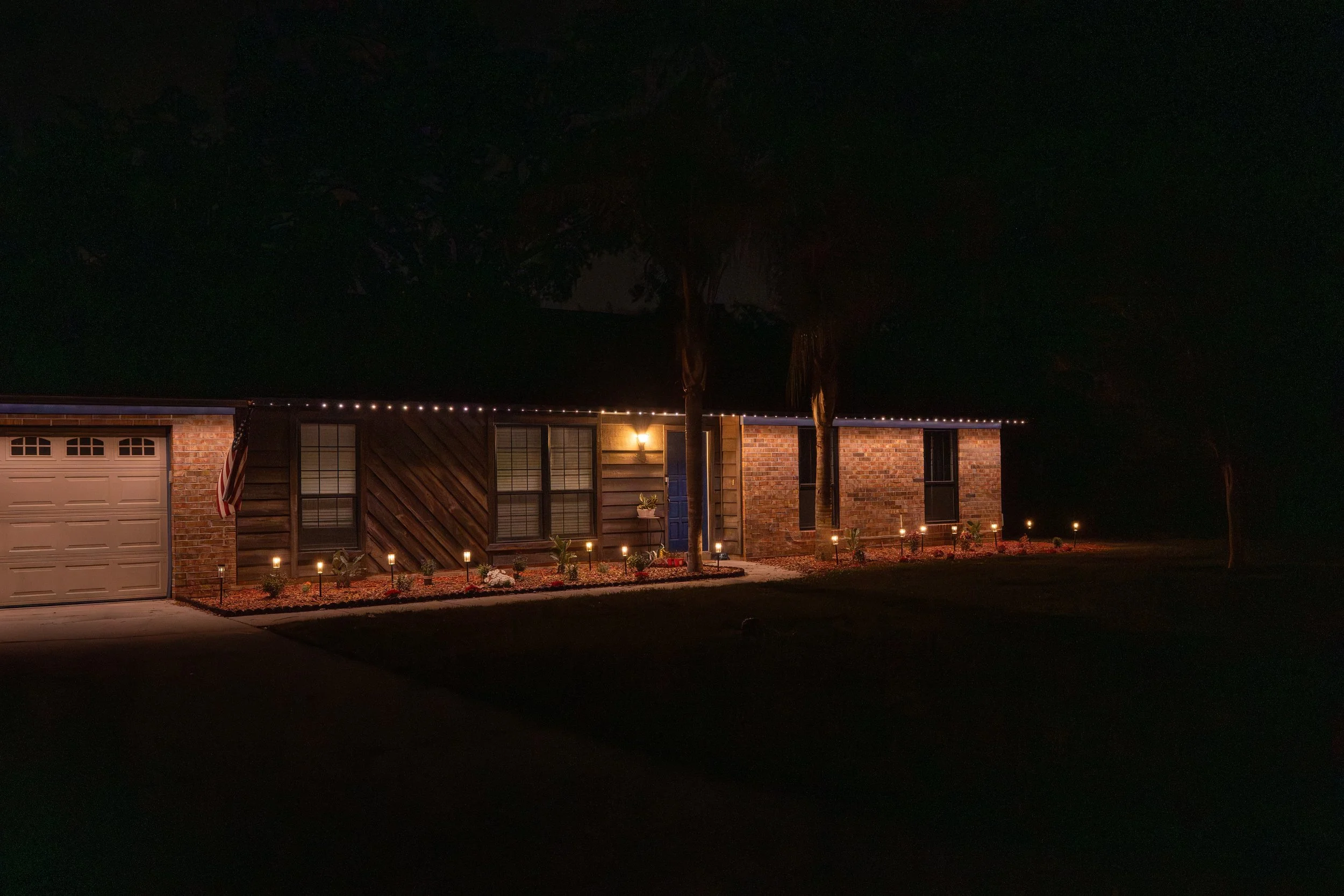 A house at night with outdoor lighting, including string lights along the roof and small garden lights illuminating a landscaped front yard.