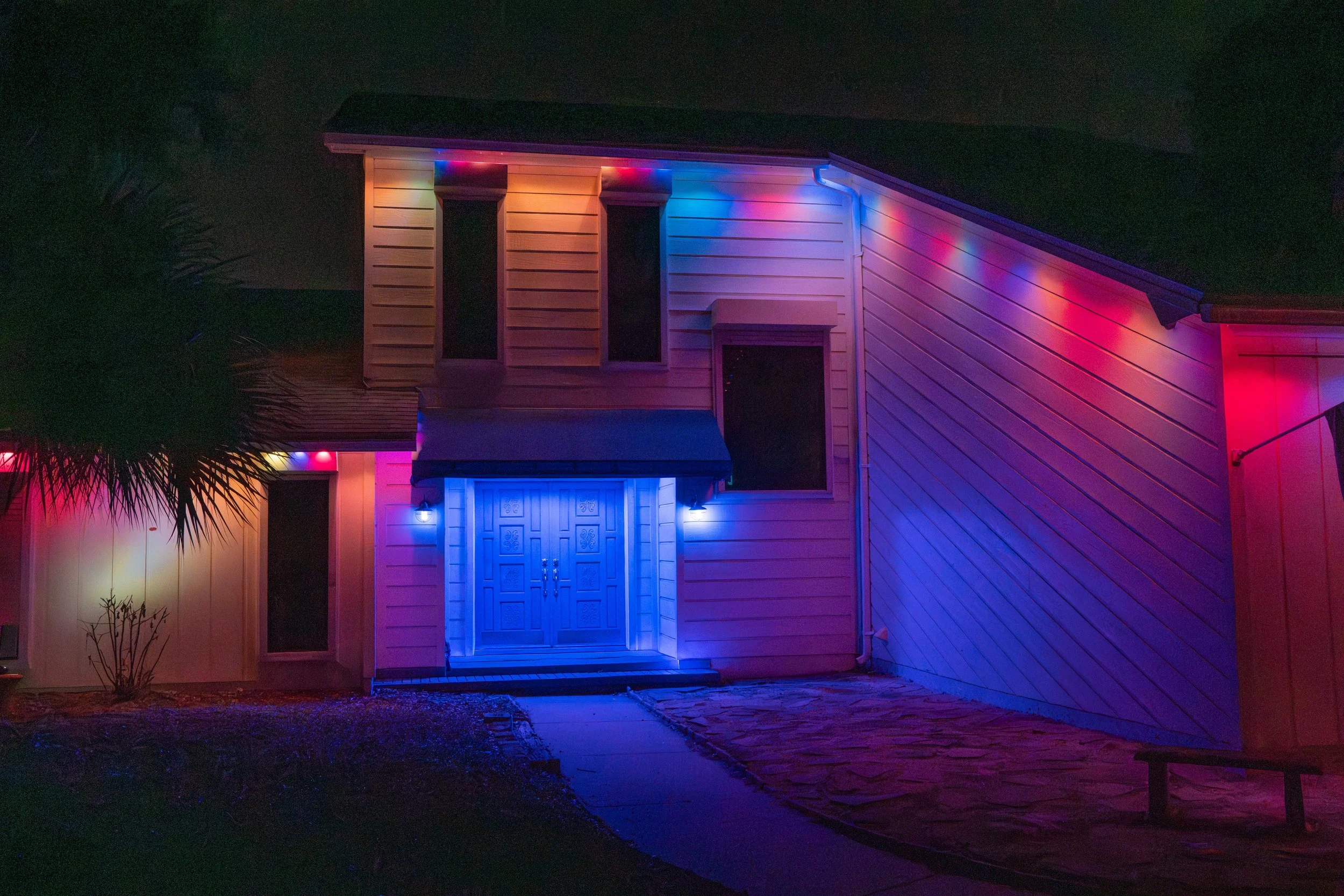A two-story house illuminated with colorful Christmas lights at night, featuring a blue front door and a stone pathway leading to the entrance.