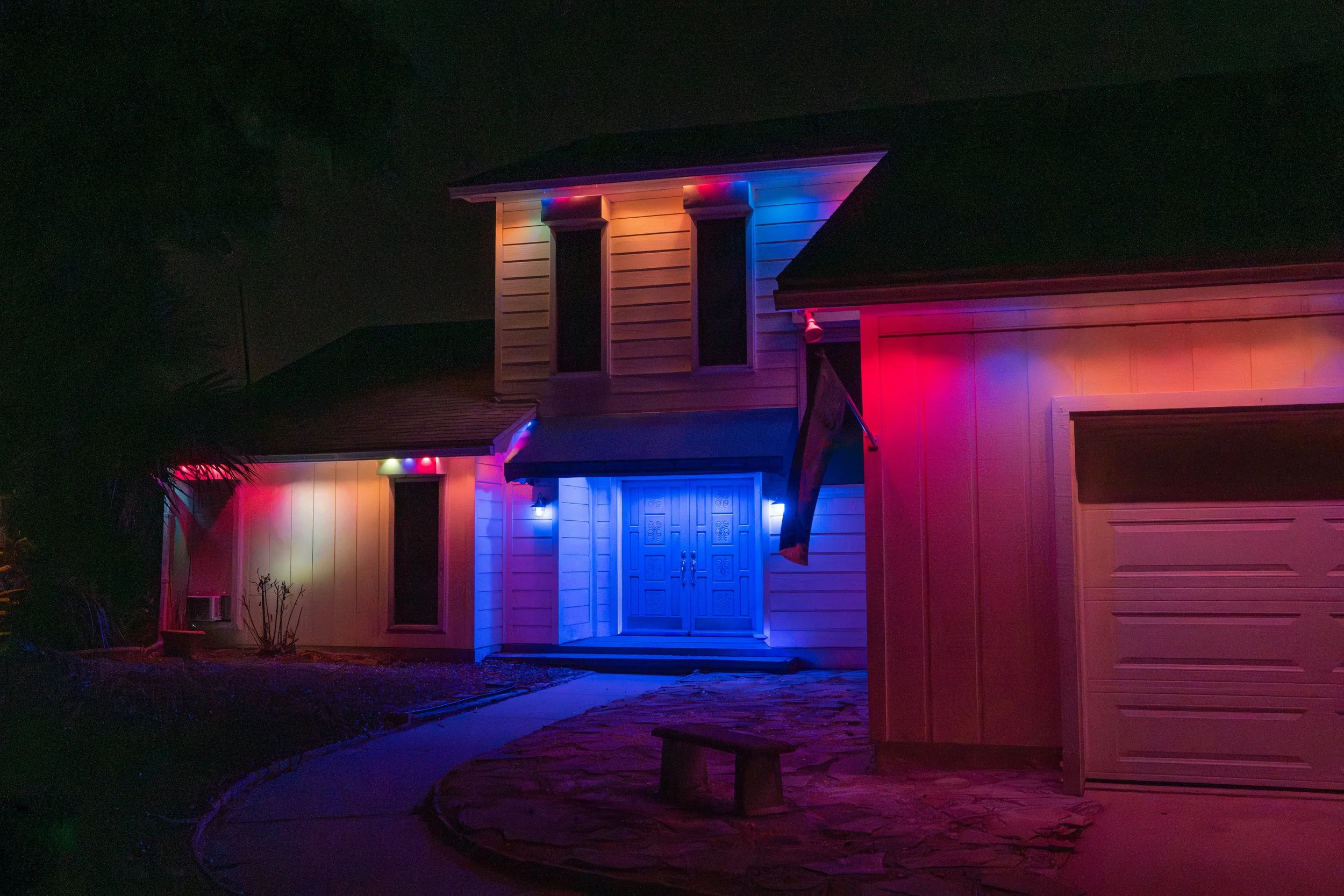 A house at night decorated with multicolored holiday lights, including blue, red, and purple, around windows, door, and roof edges.