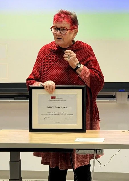 An older woman with short red hair and glasses holding a framed certificate at a presentation or award ceremony.