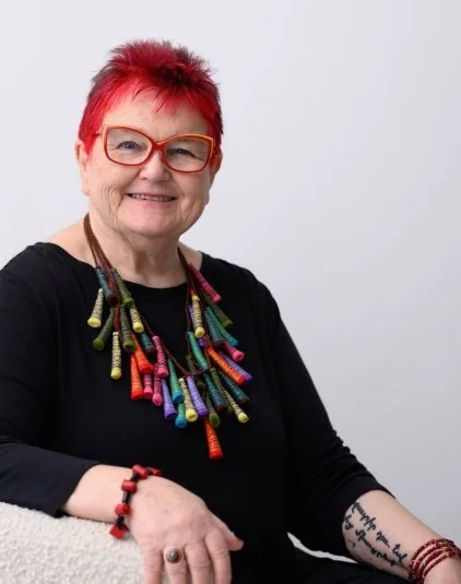 A smiling woman with short red hair and red glasses, wearing a black top and colorful beaded necklace, sitting against a plain light background.