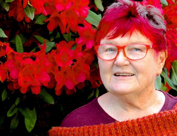 An older woman with short red and purple hair, wearing red glasses, standing in front of red and pink flowers.