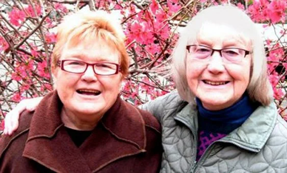 Two older women smiling outdoors in front of pink flowering trees.