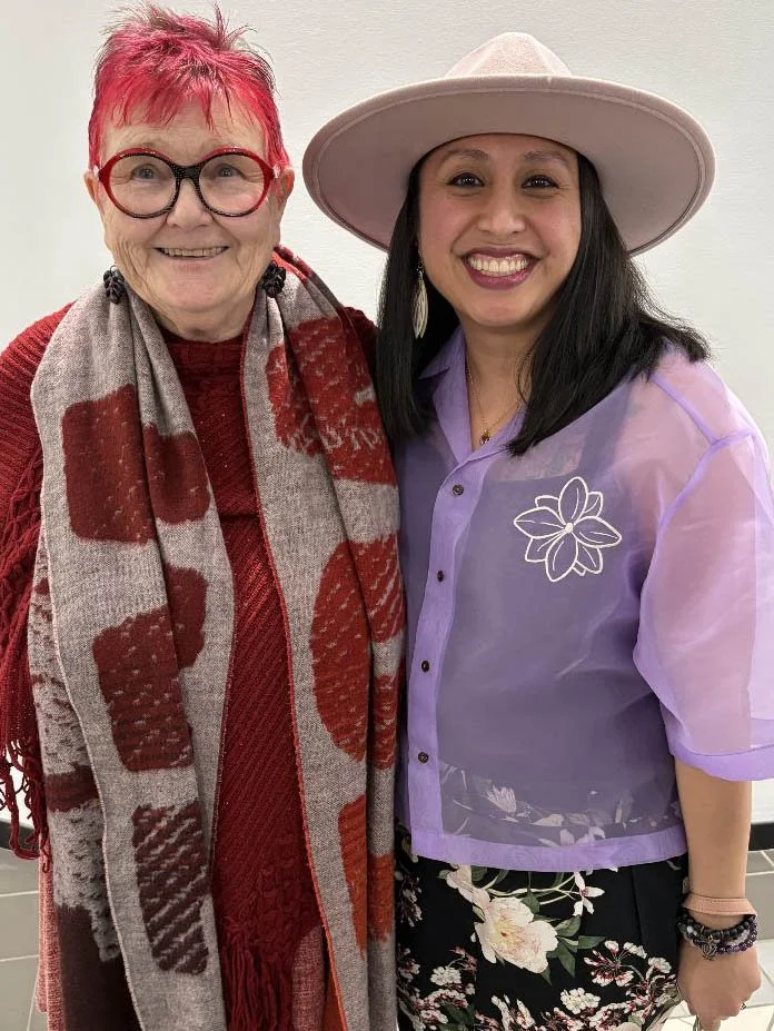 Two women smiling and standing close together, one with short pink hair, glasses, and a red sweater paired with a patterned scarf, the other with long black hair, wearing a wide-brimmed hat, lavender jacket with a floral design, and floral skirt.