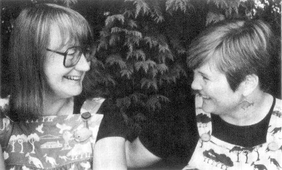 Two women smiling at each other outdoors, with trees in the background.