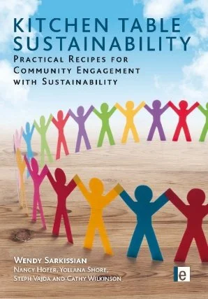 Book cover titled "Kitchen Table Sustainability" with colorful paper chain figures holding hands in a circle, set on a wooden table background.