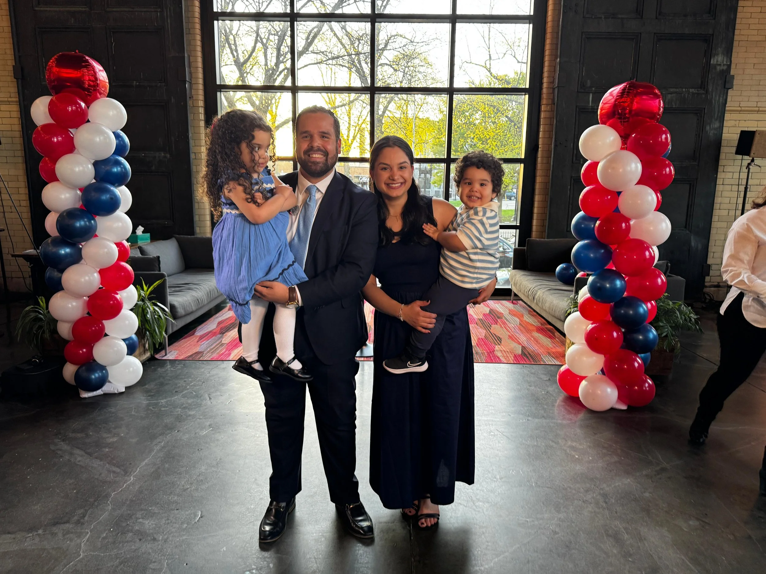 A group of four people, two children and two adults, posing together at a celebration with red, white, and blue balloon columns in the background.