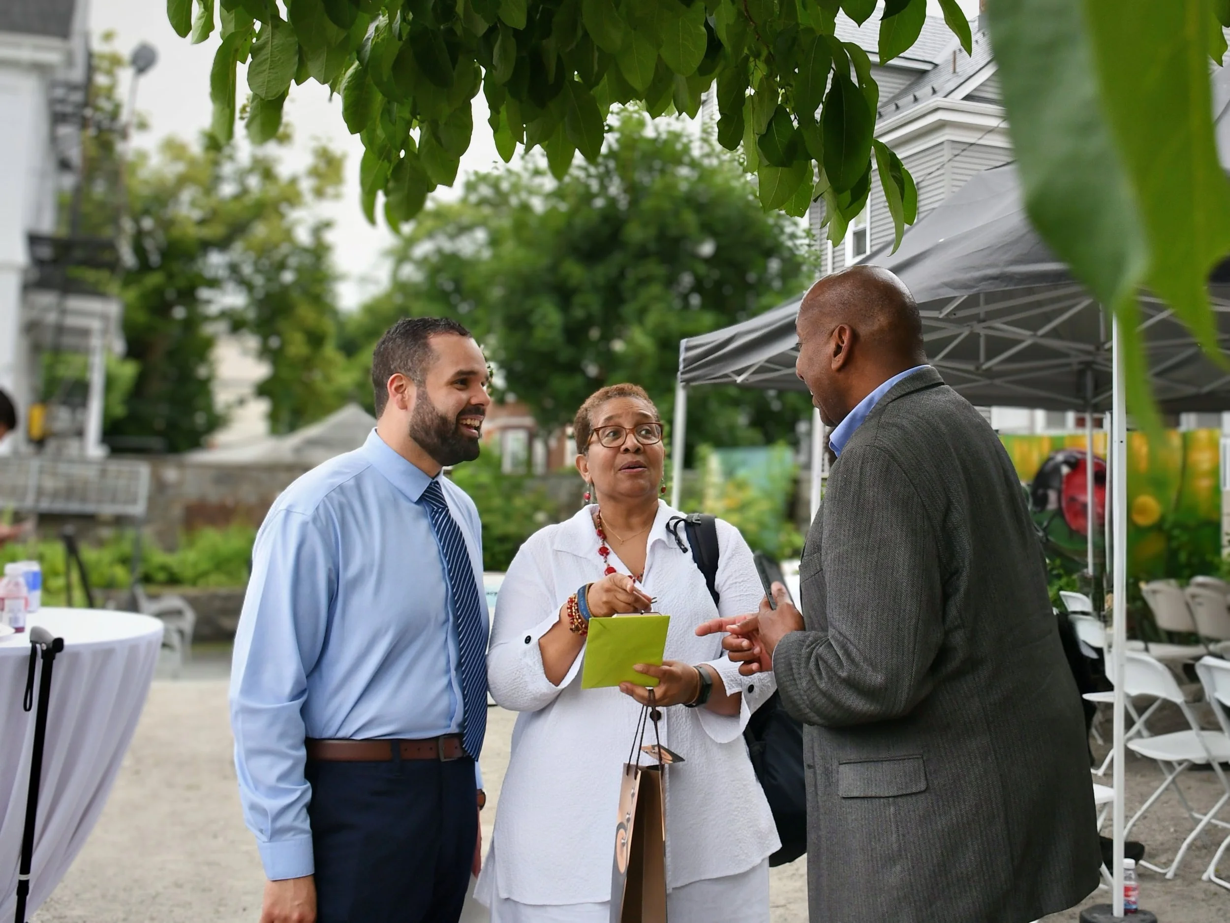 Three people talking at an outdoor event, with tables and tents in the background, green trees overhead, and a building visible behind them.