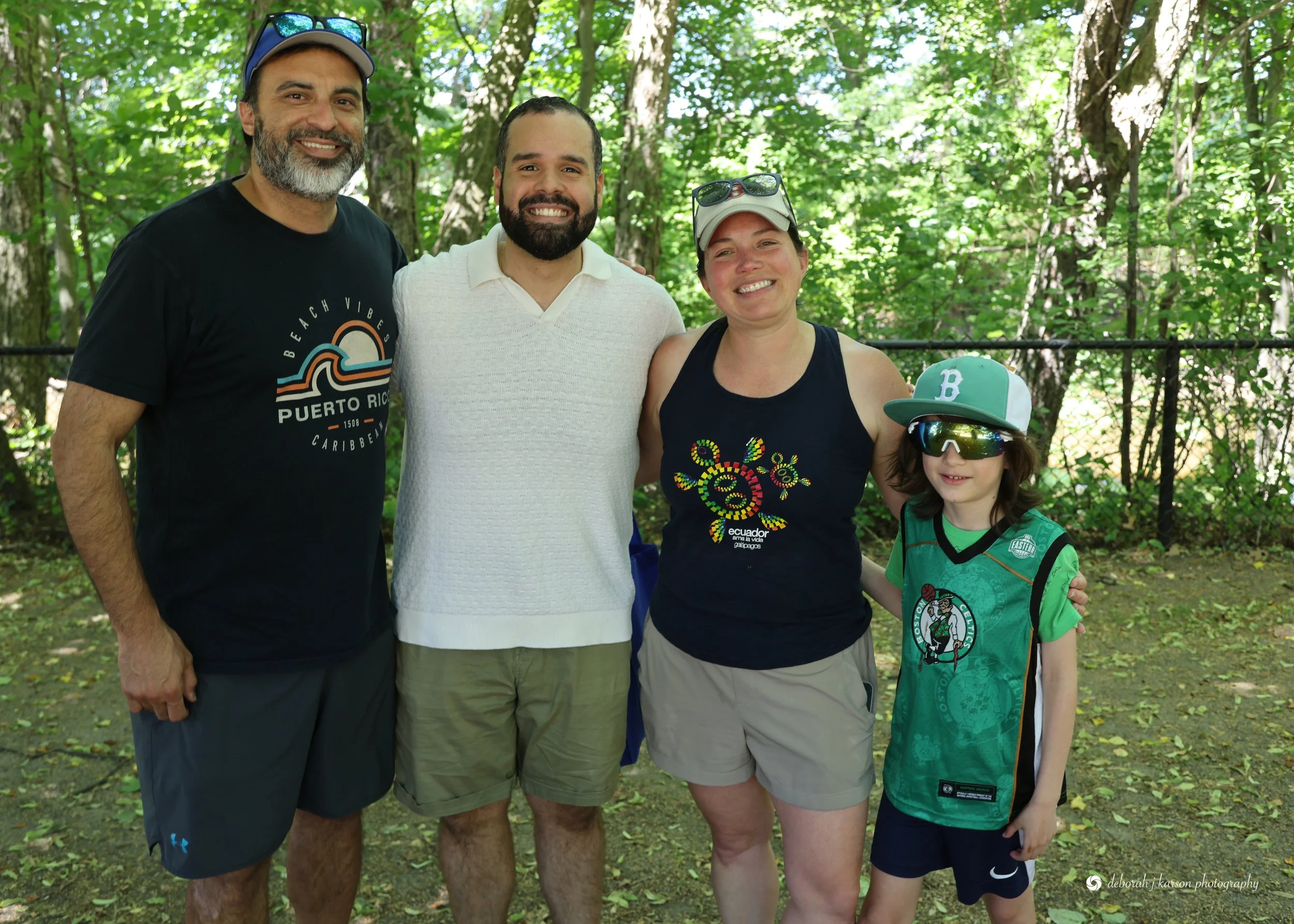 A group of four people standing outdoors in a wooded area, smiling at the camera. The group includes three adults and one child, all dressed casually for a nature outing.