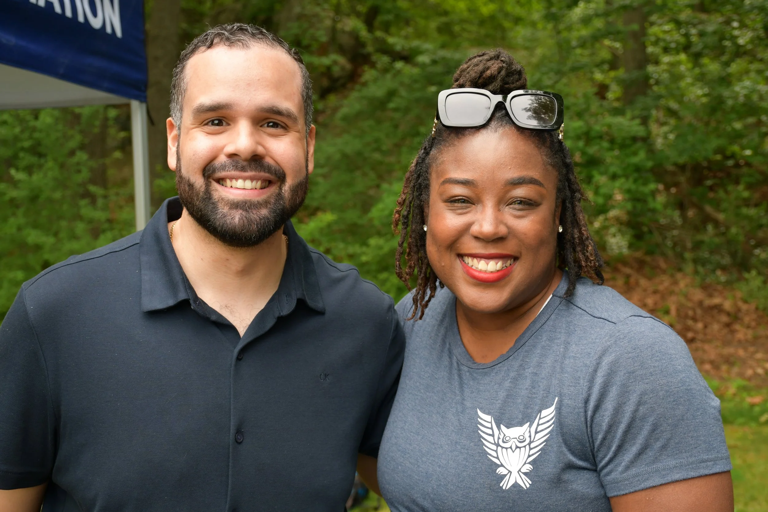 A man and a woman smiling outdoors, surrounded by green trees. The woman has sunglasses on her head and is wearing a gray t-shirt with a white owl graphic, while the man is wearing a dark shirt.