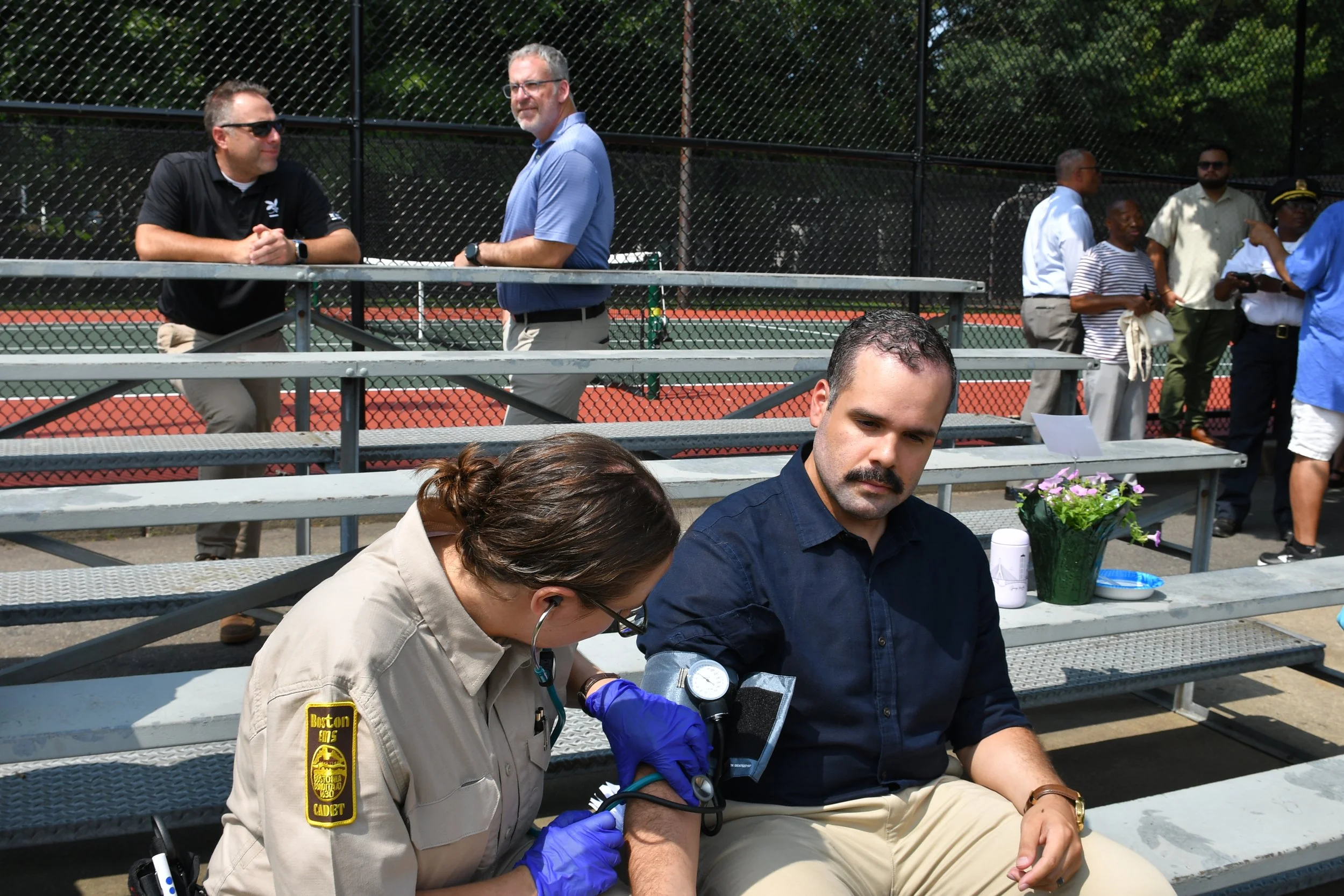 A woman in a beige uniform with a badge is administering a vaccine to a man in a blue shirt sitting on a bench, while a nurse wearing purple gloves is holding his arm. In the background, several people are standing near a chain-link fence at an outdoor tennis court.
